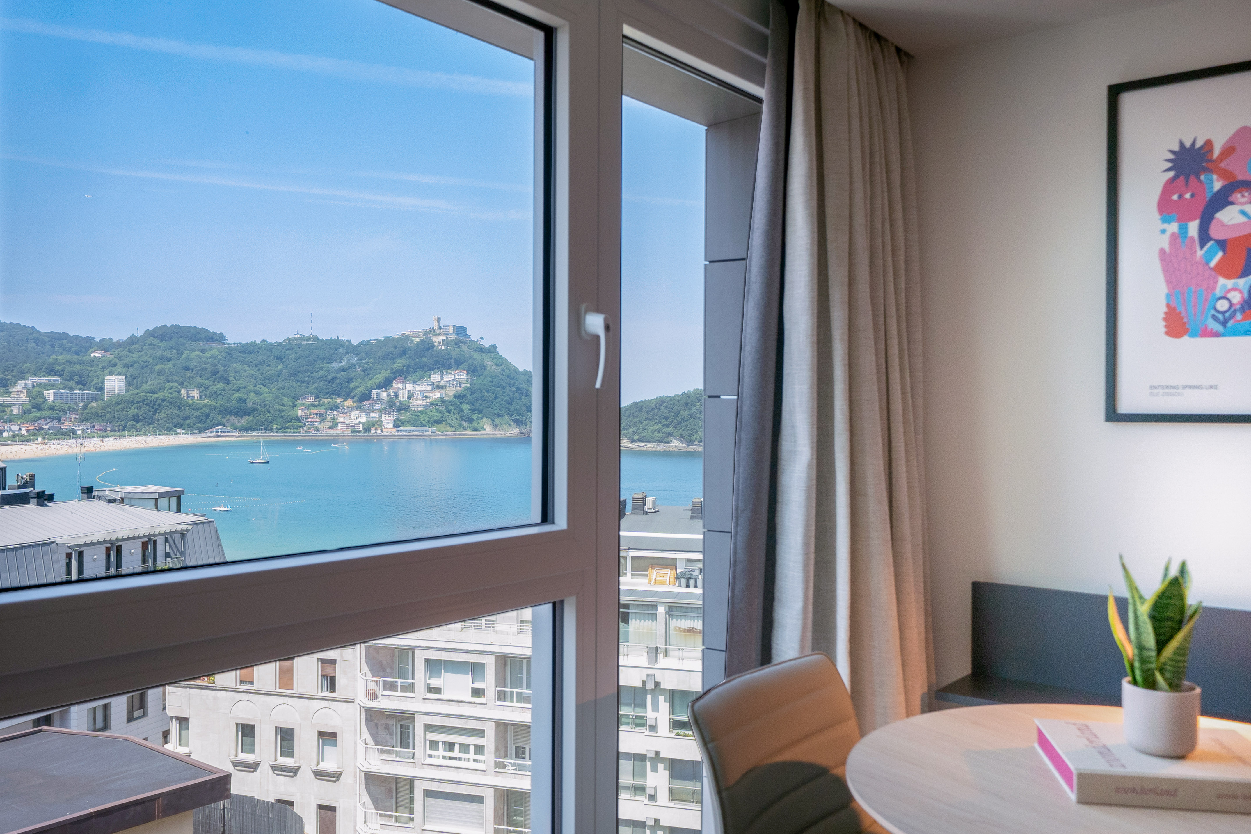 Vista desde ventana a la Bahía de La Concha, San Sebastián, con playa y montañas.