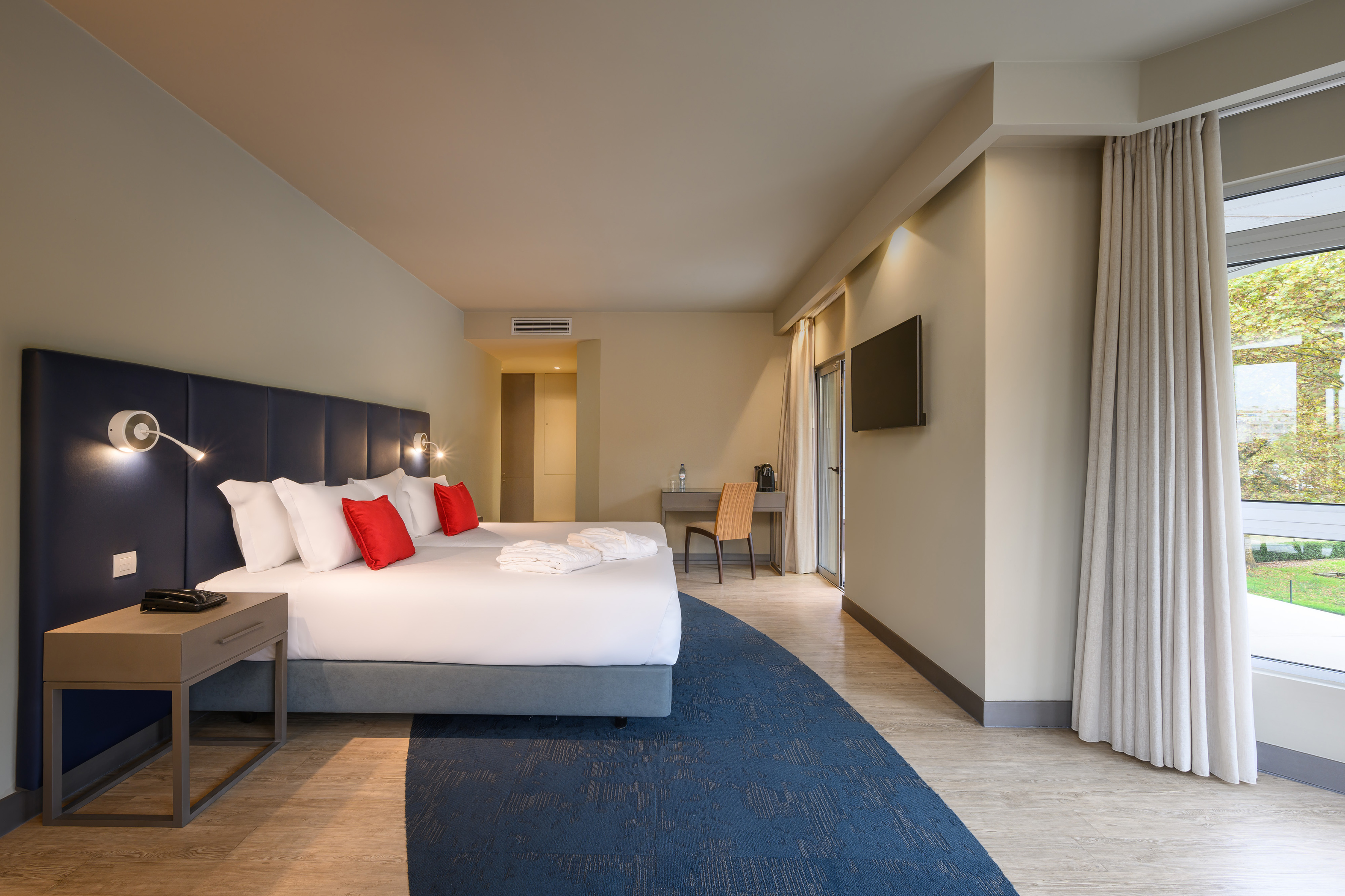 Modern hotel room with king bed, red pillows, desk, TV, and window view.