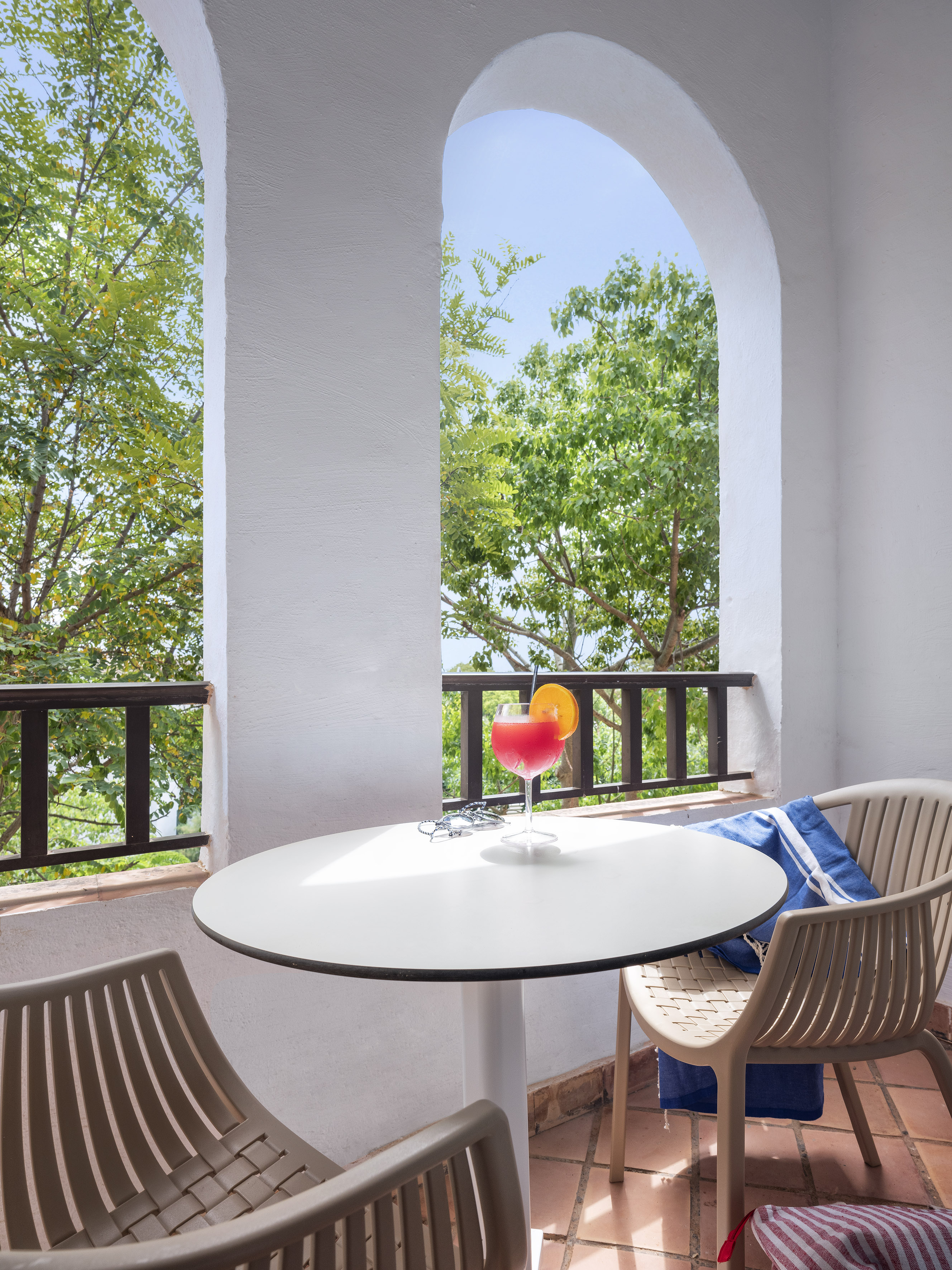 Arched balcony with red cocktail, outdoor chairs, and lush green tree view.