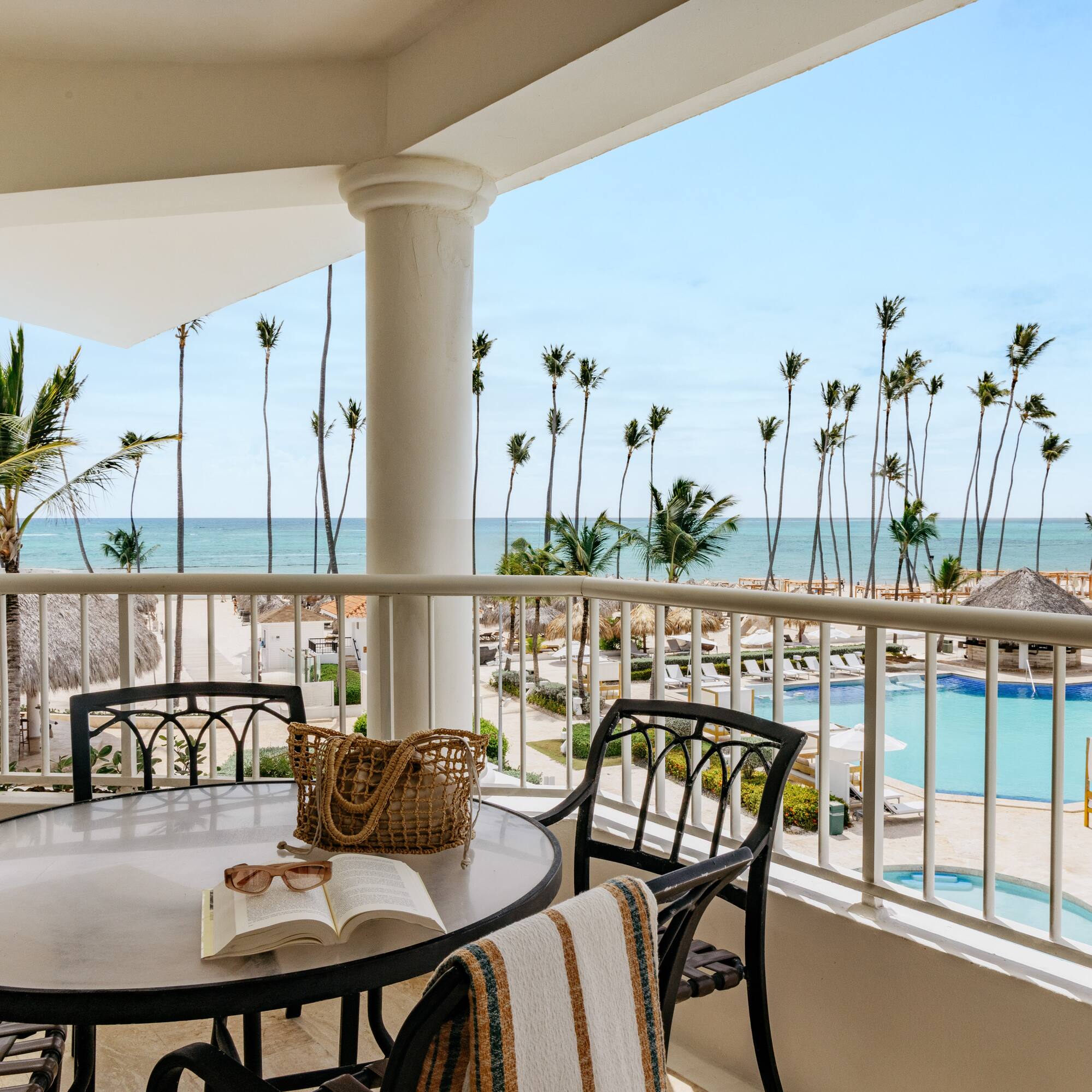 Oceanfront balcony overlooking tropical beach, palm trees, and resort pool.