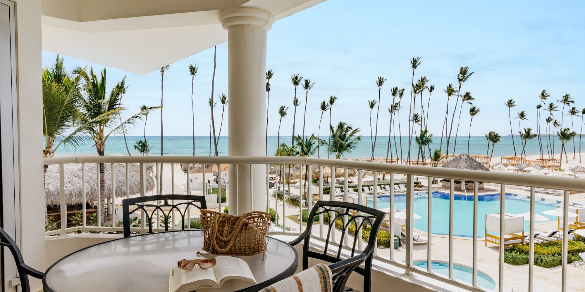 Oceanfront balcony overlooking tropical beach, palm trees, and resort pool.