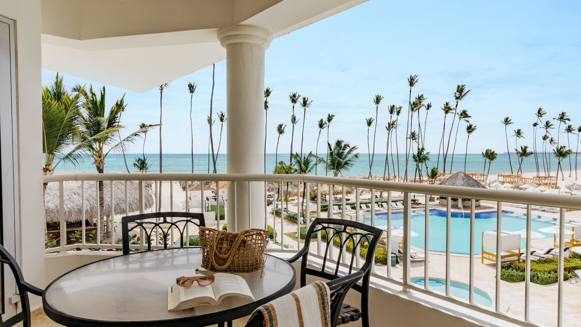 Oceanfront balcony overlooking tropical beach, palm trees, and resort pool.