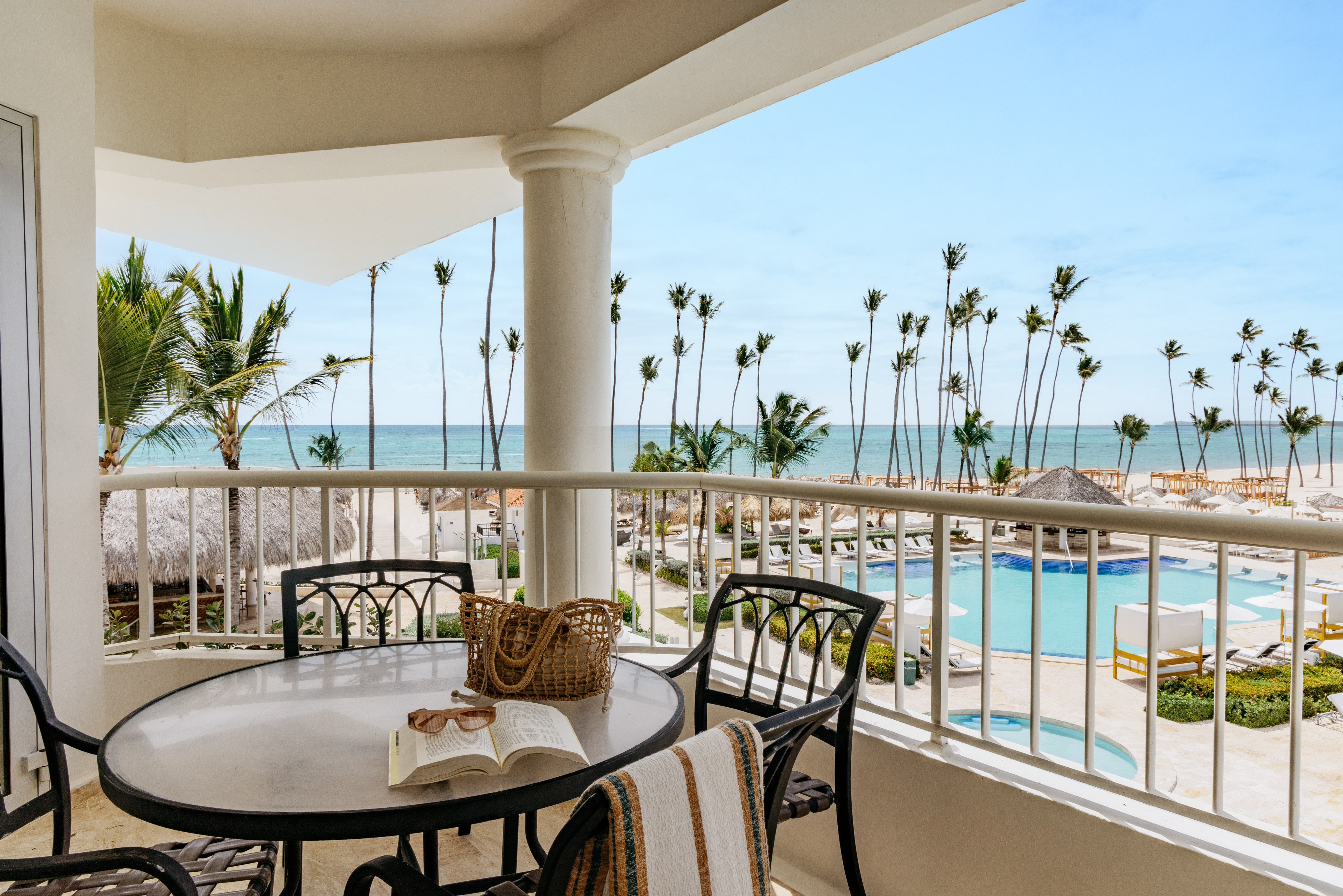 Oceanfront balcony overlooking tropical beach, palm trees, and resort pool.