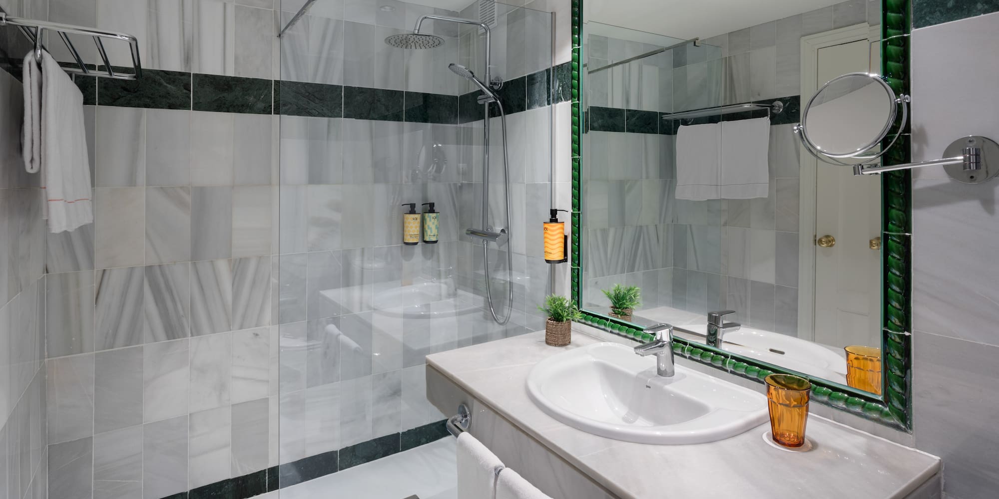 Modern bathroom with white marble tiles, glass shower, and green-framed mirror.