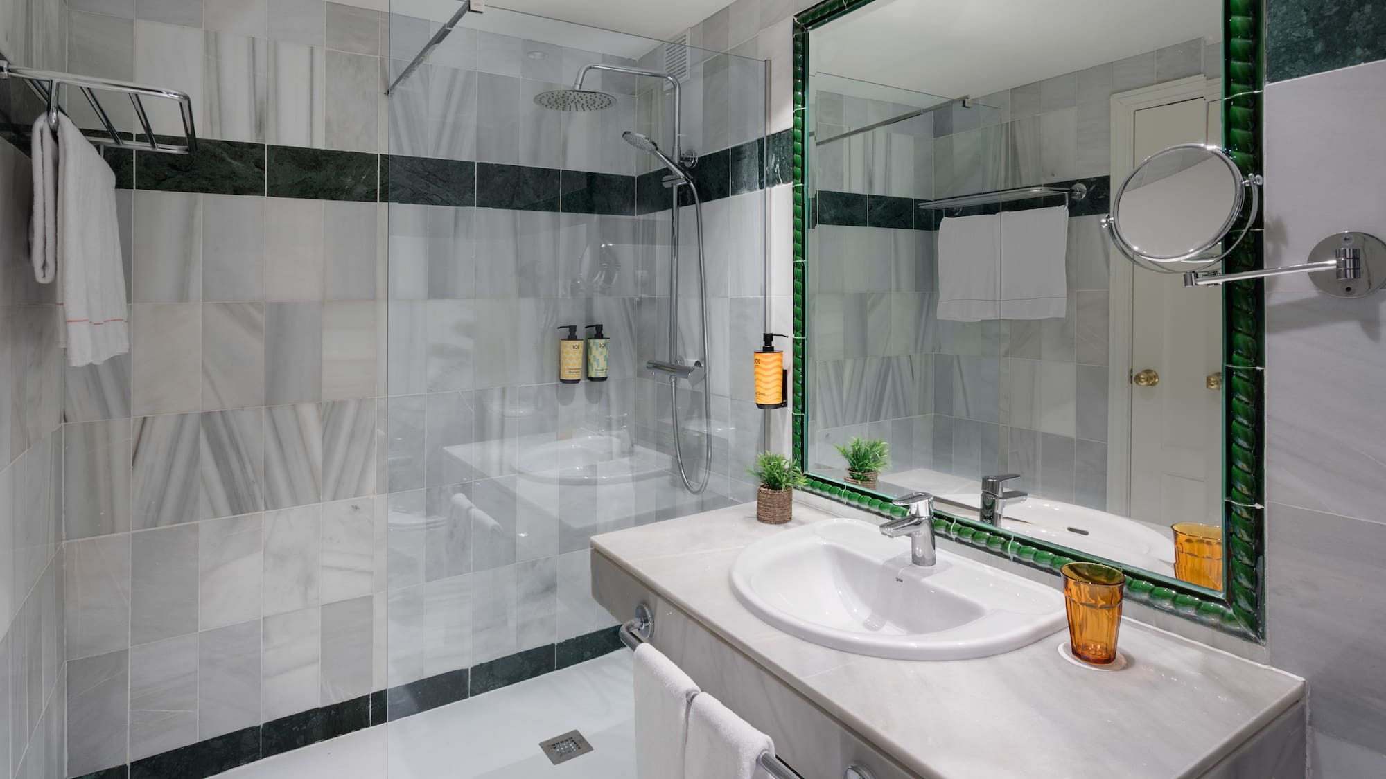 Baño hotel con ducha de mármol gris, lavabo moderno y espejo verde.