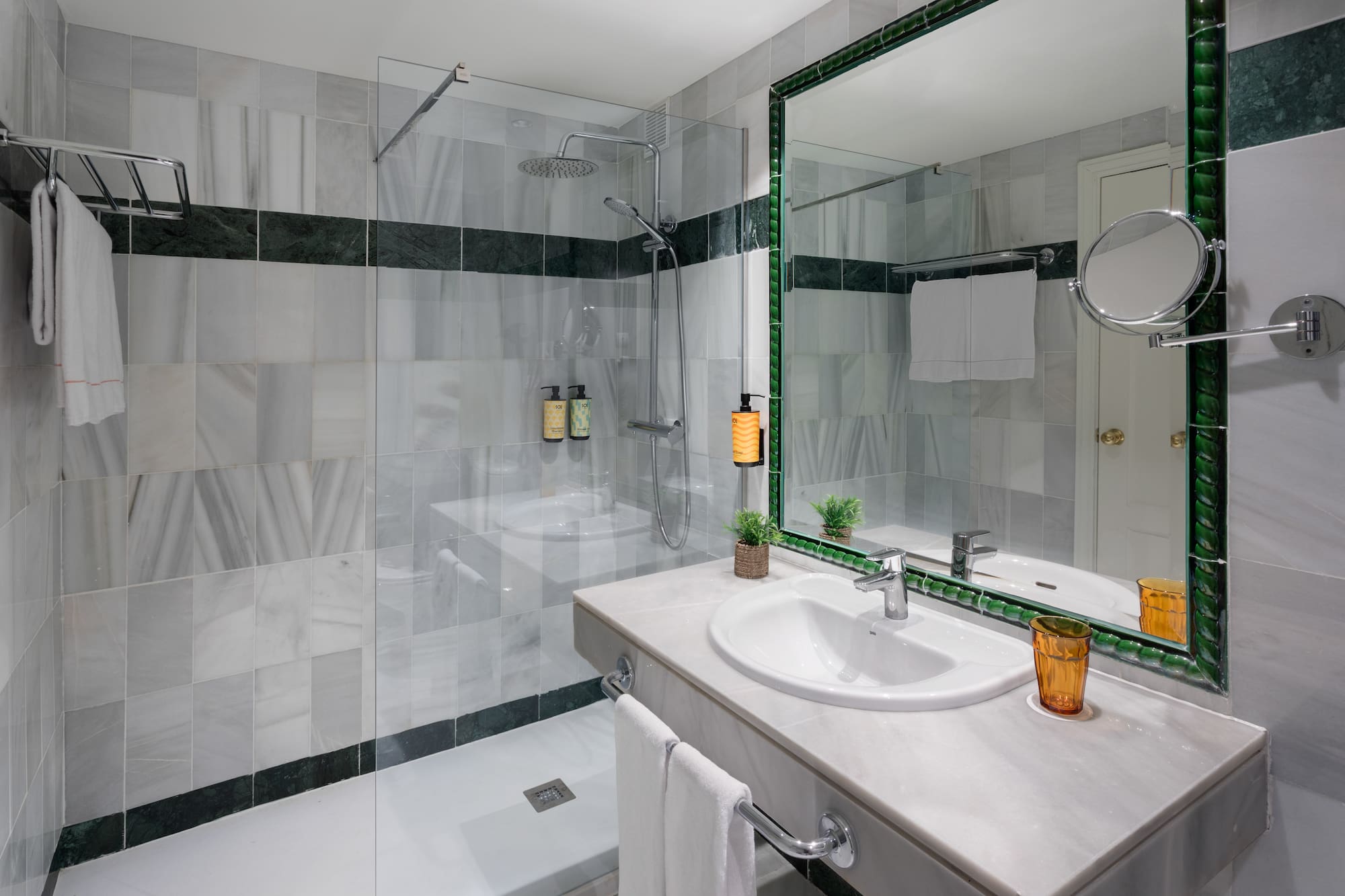 Baño hotel con ducha de mármol gris, lavabo moderno y espejo verde.