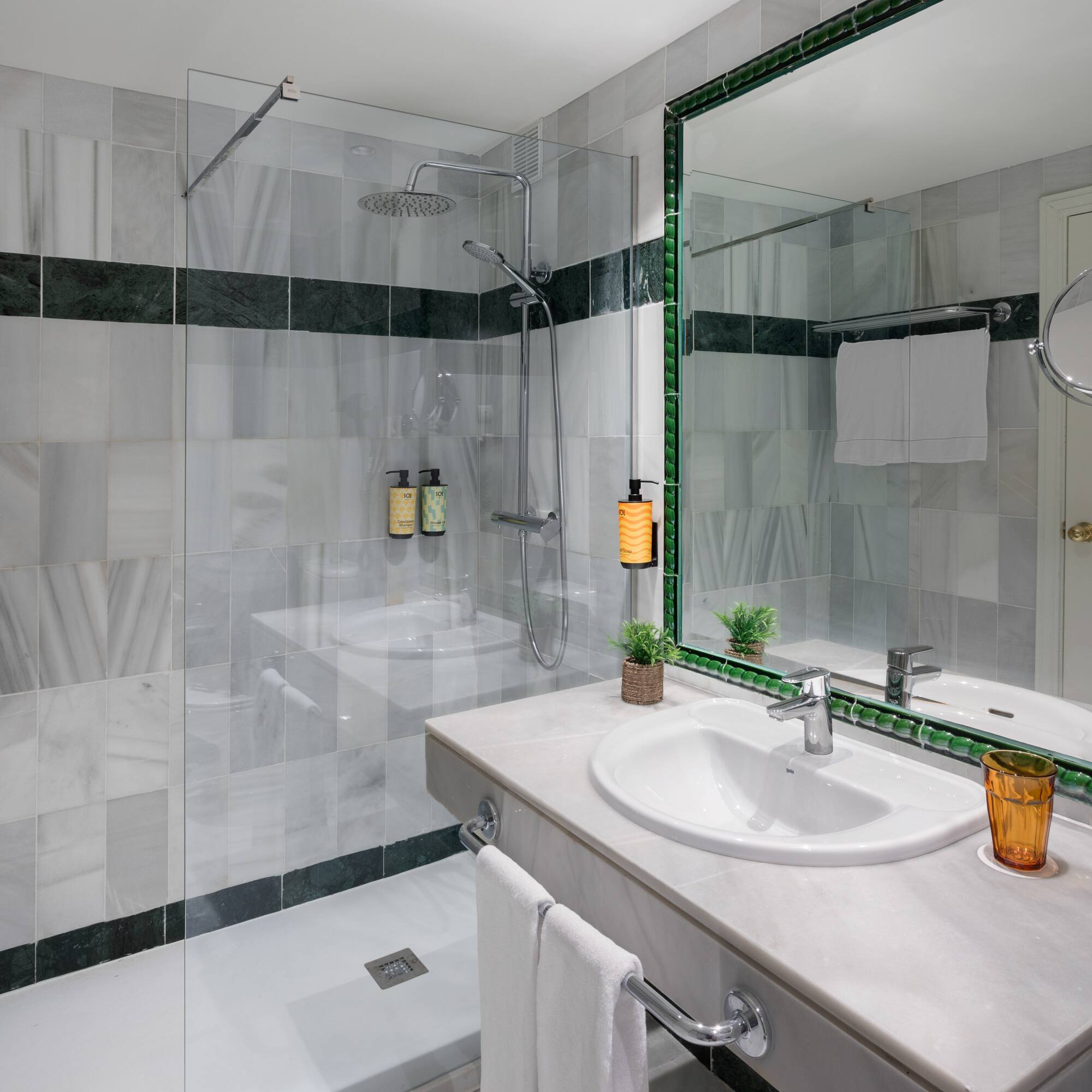 Baño hotel con ducha de mármol gris, lavabo moderno y espejo verde.