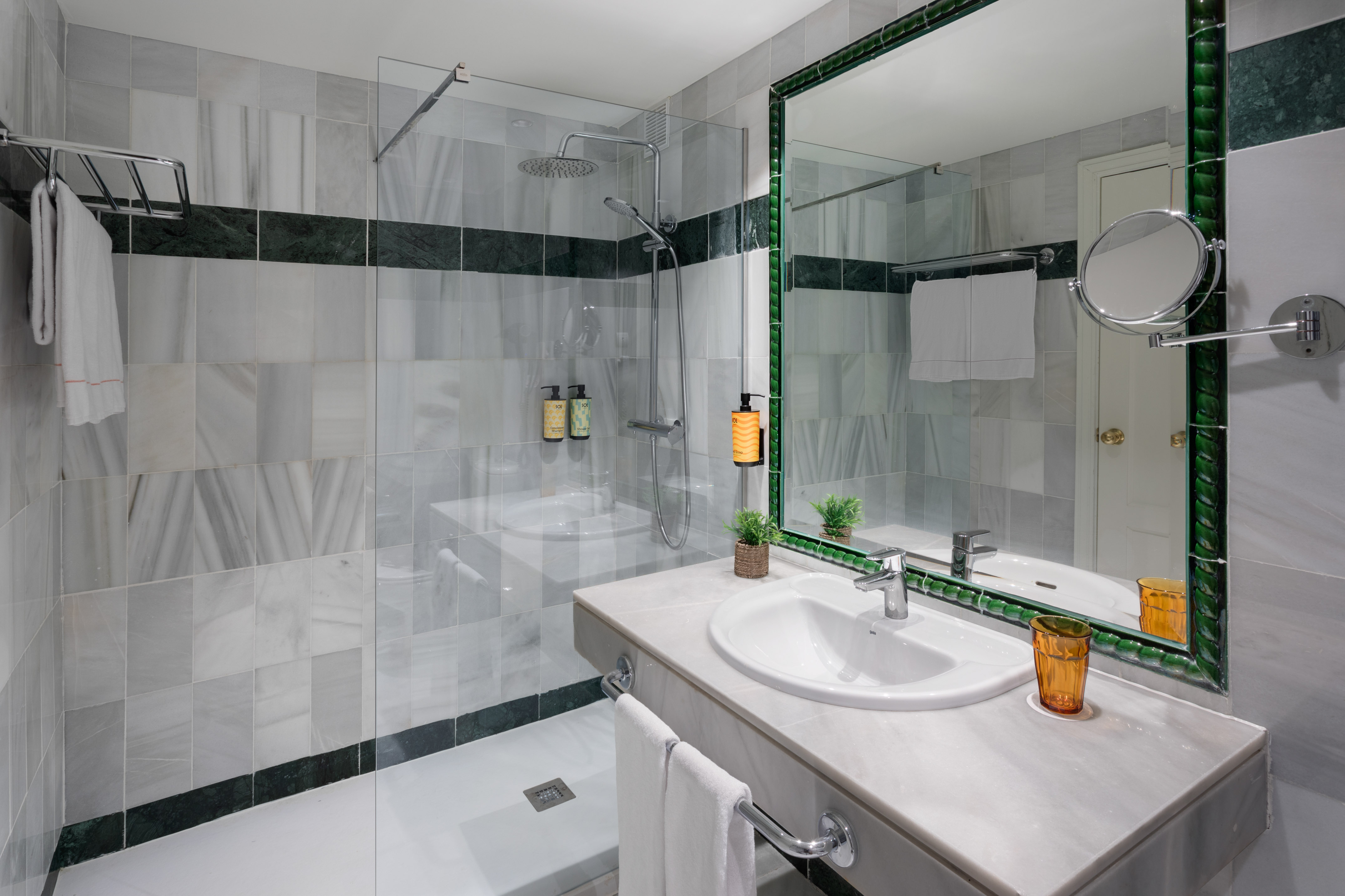 Modern bathroom with white marble tiles, glass shower, and green-framed mirror.