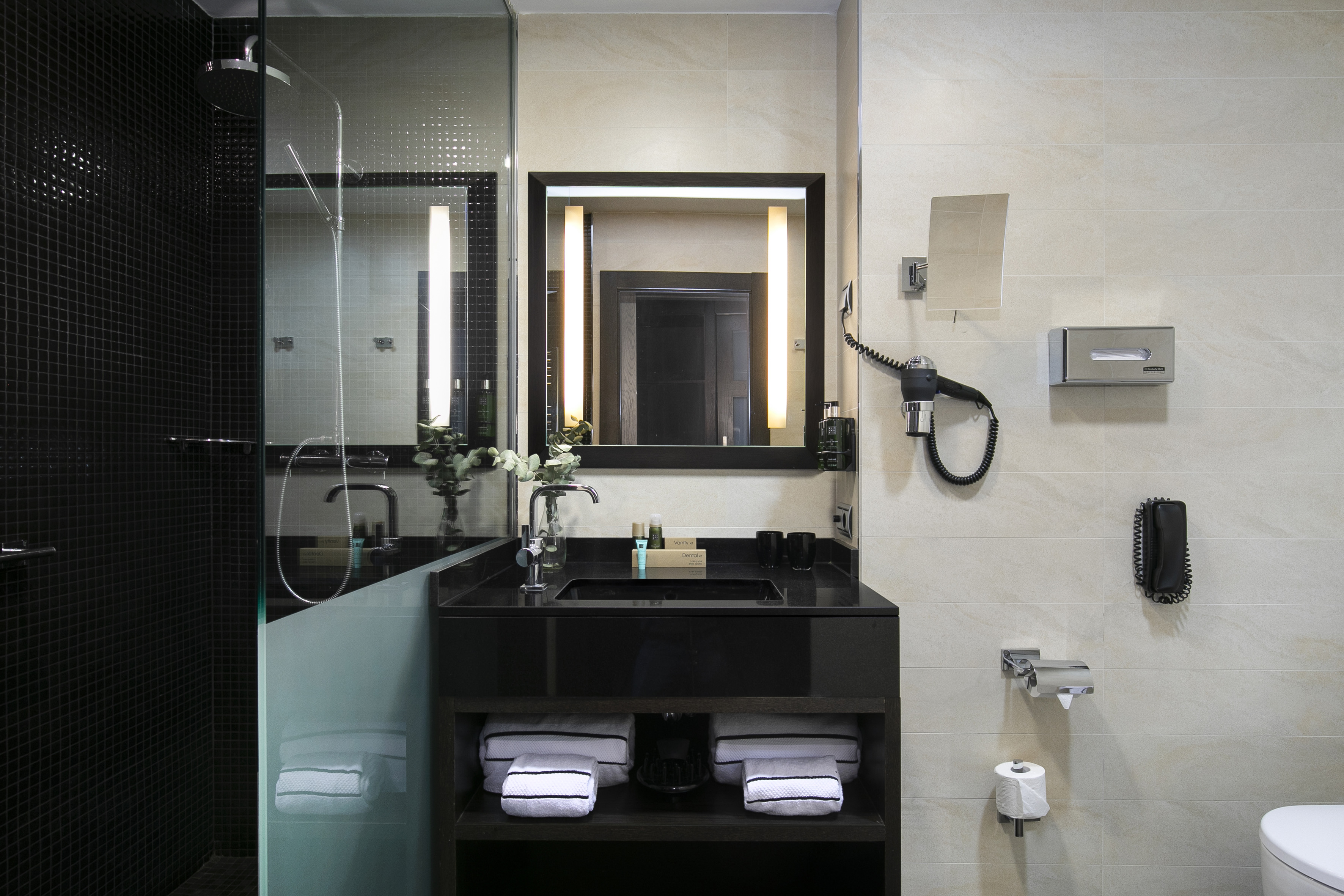 Modern hotel bathroom with black vanity, illuminated mirror, and glass shower.