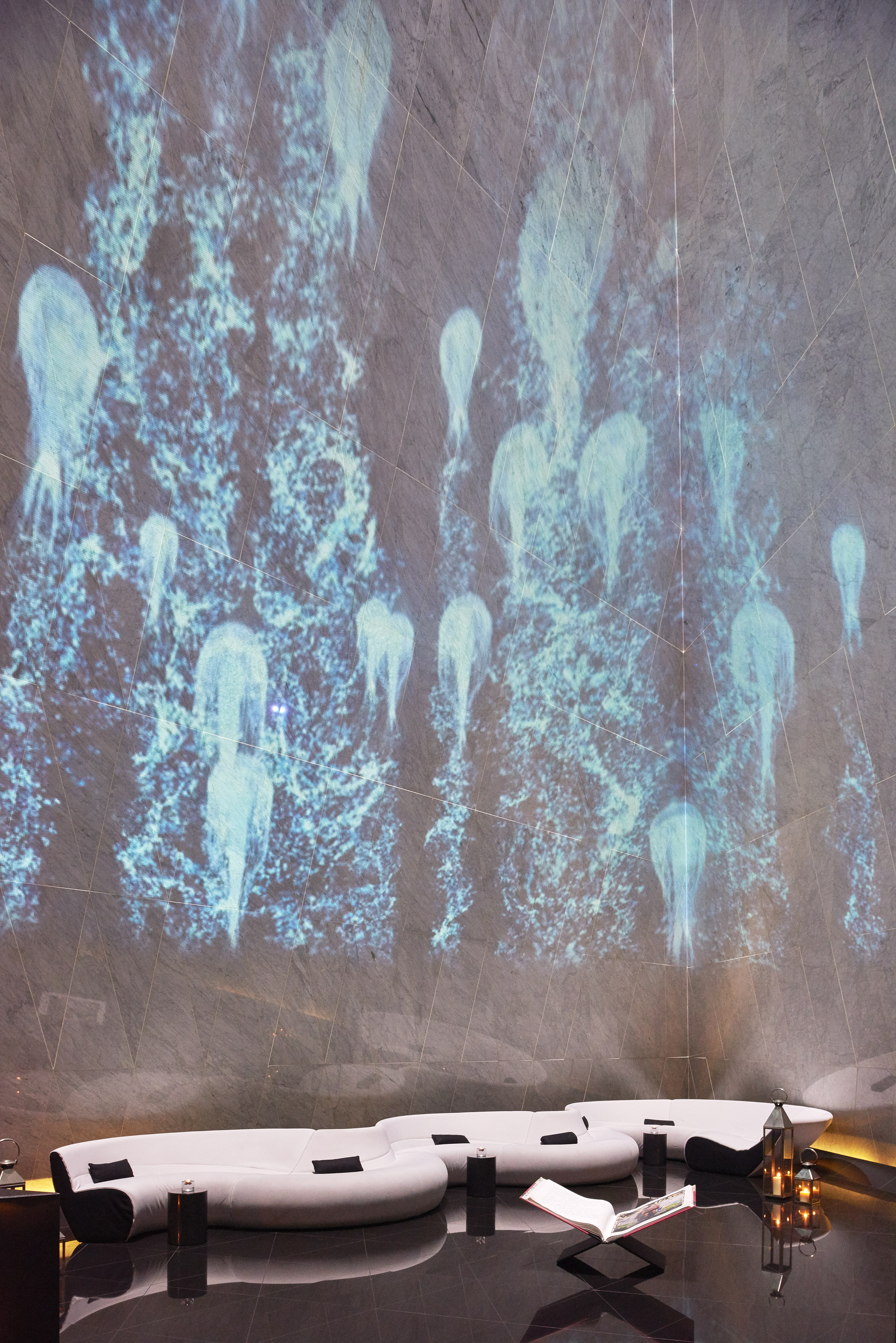 Modern lounge with blue jellyfish projection art on stone walls, curved white sofas.