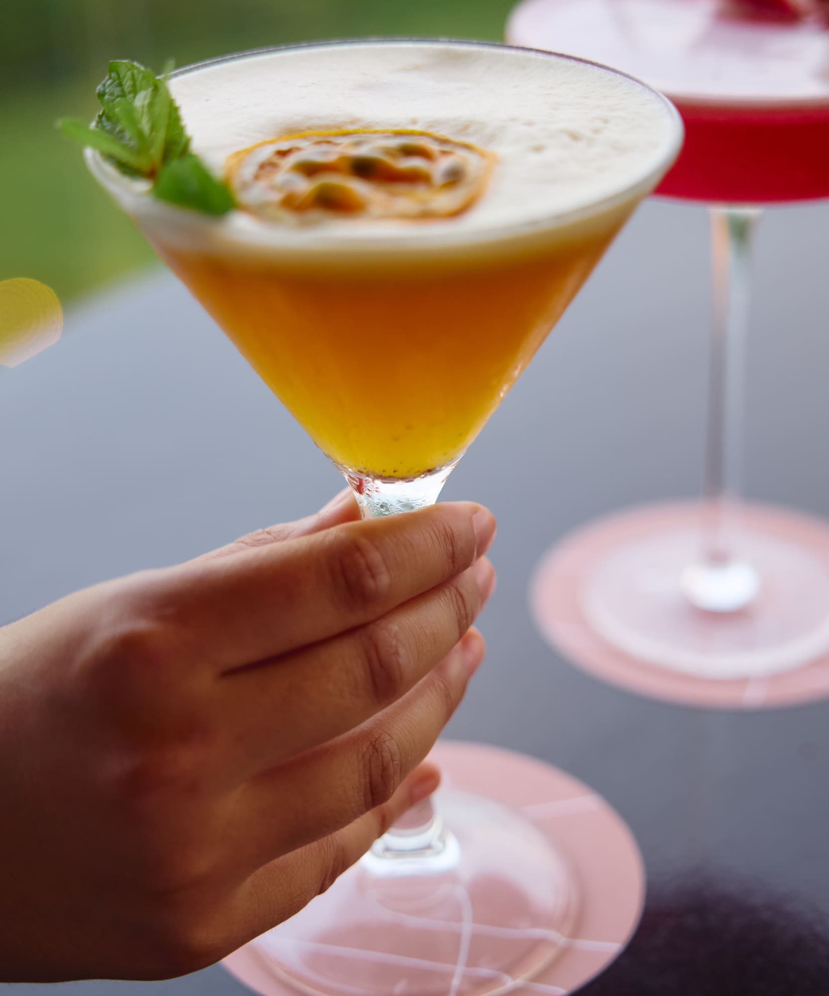 Hand holding a passion fruit martini with mint and foam. Another cocktail and candle in background.