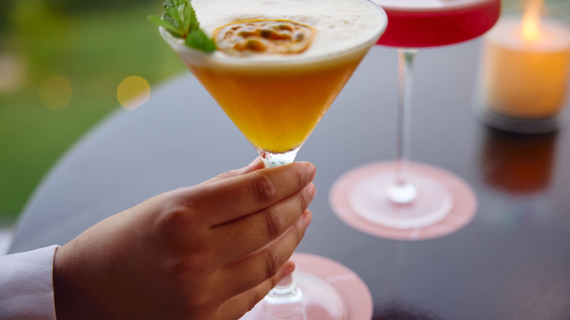 Hand holds passion fruit martini with mint. Red cocktail, candle.