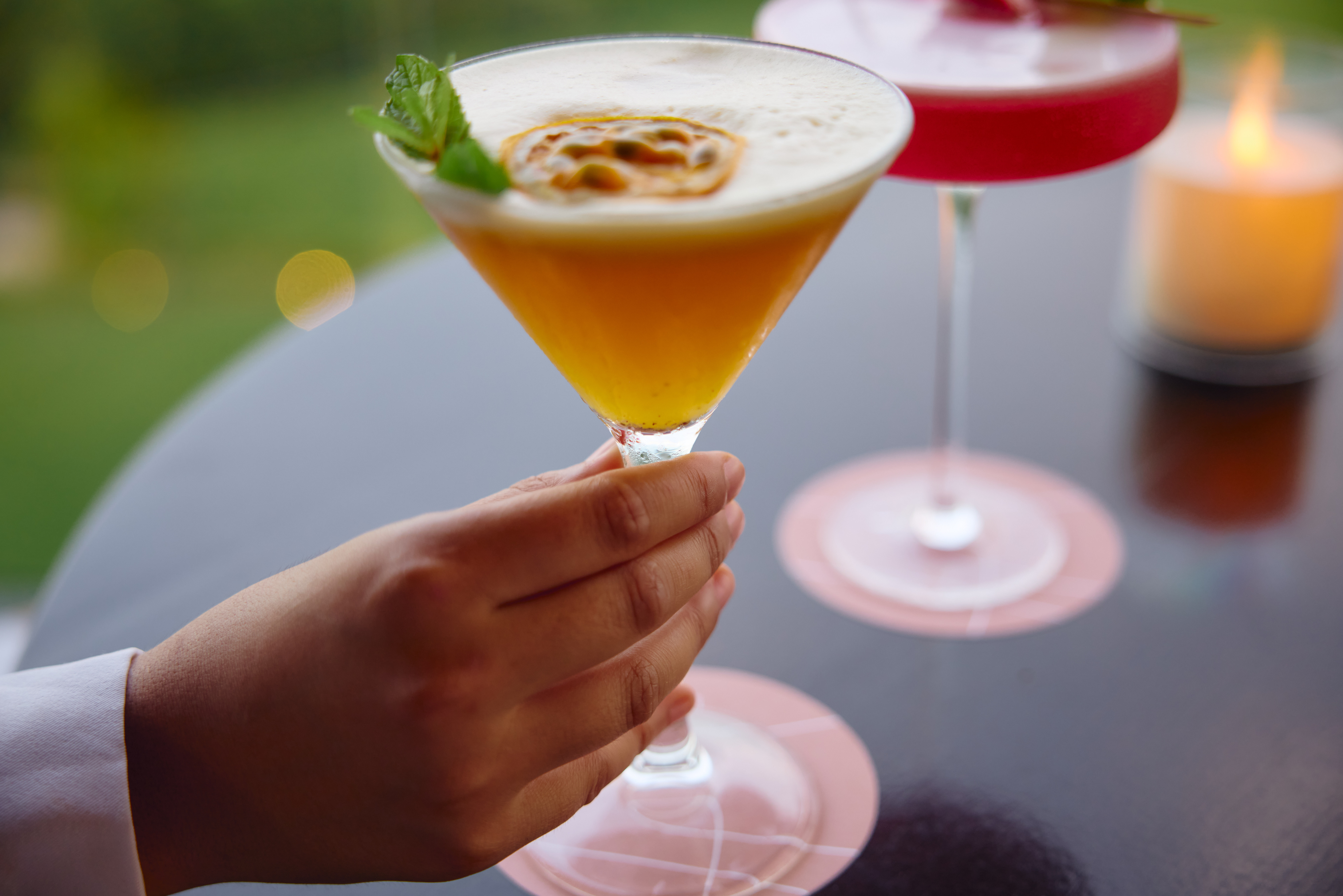 Hand holding a passion fruit martini with mint and foam. Another cocktail and candle in background.