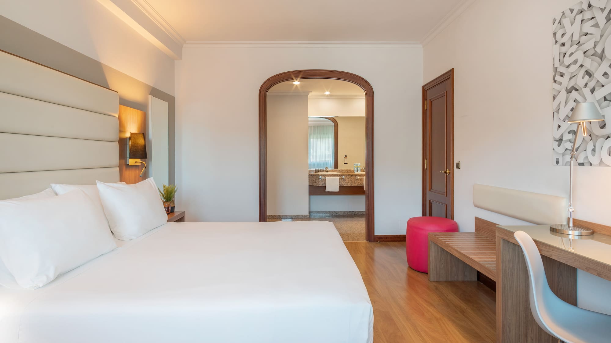 Modern hotel bedroom with white bed, desk, and arched doorway to bathroom.