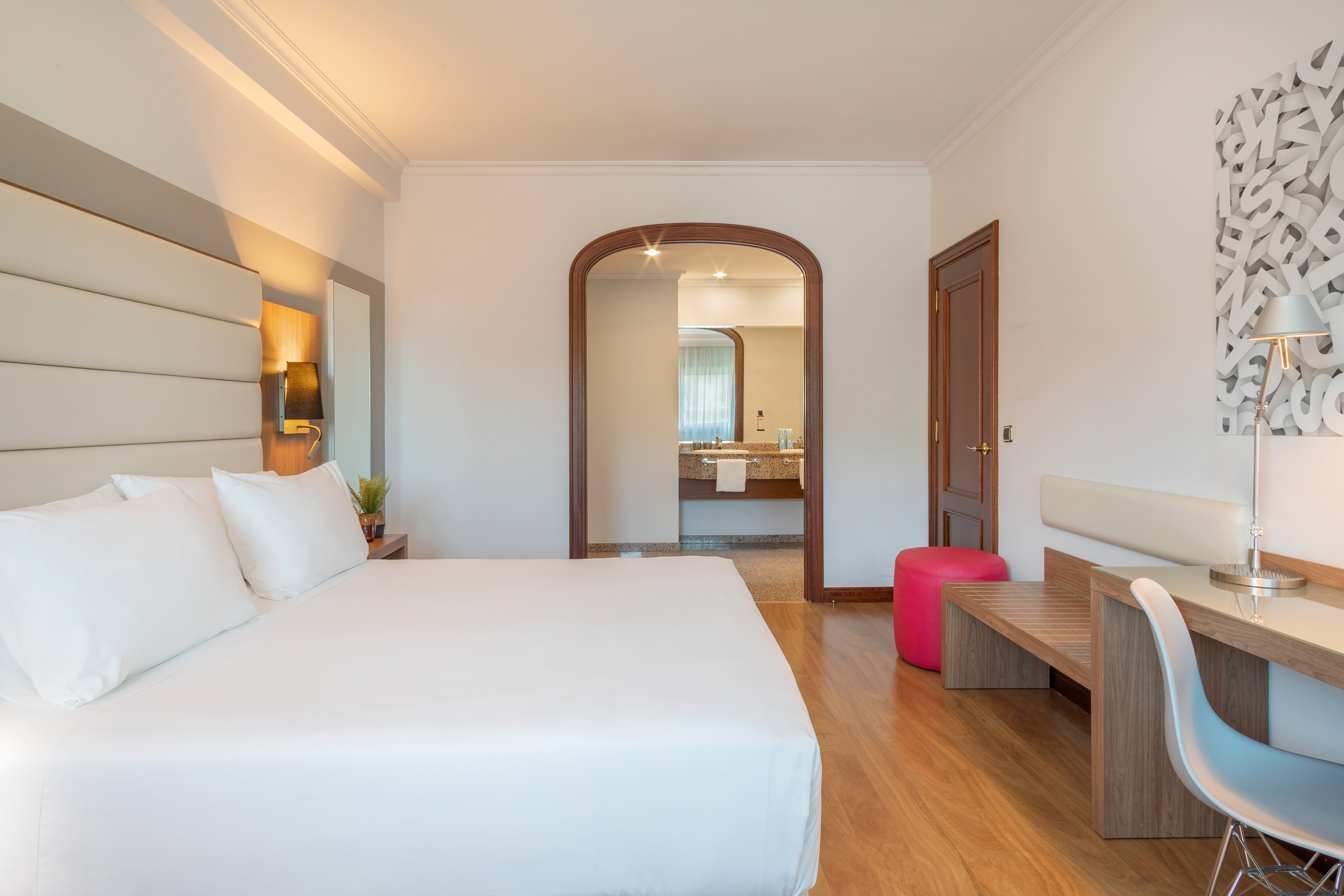 Modern hotel bedroom with white bed, desk, and arched doorway to bathroom.