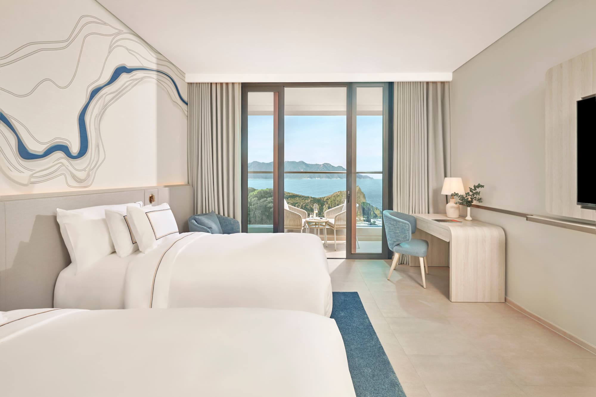 Bright hotel room with two beds, large balcony overlooking ocean and cliffs.