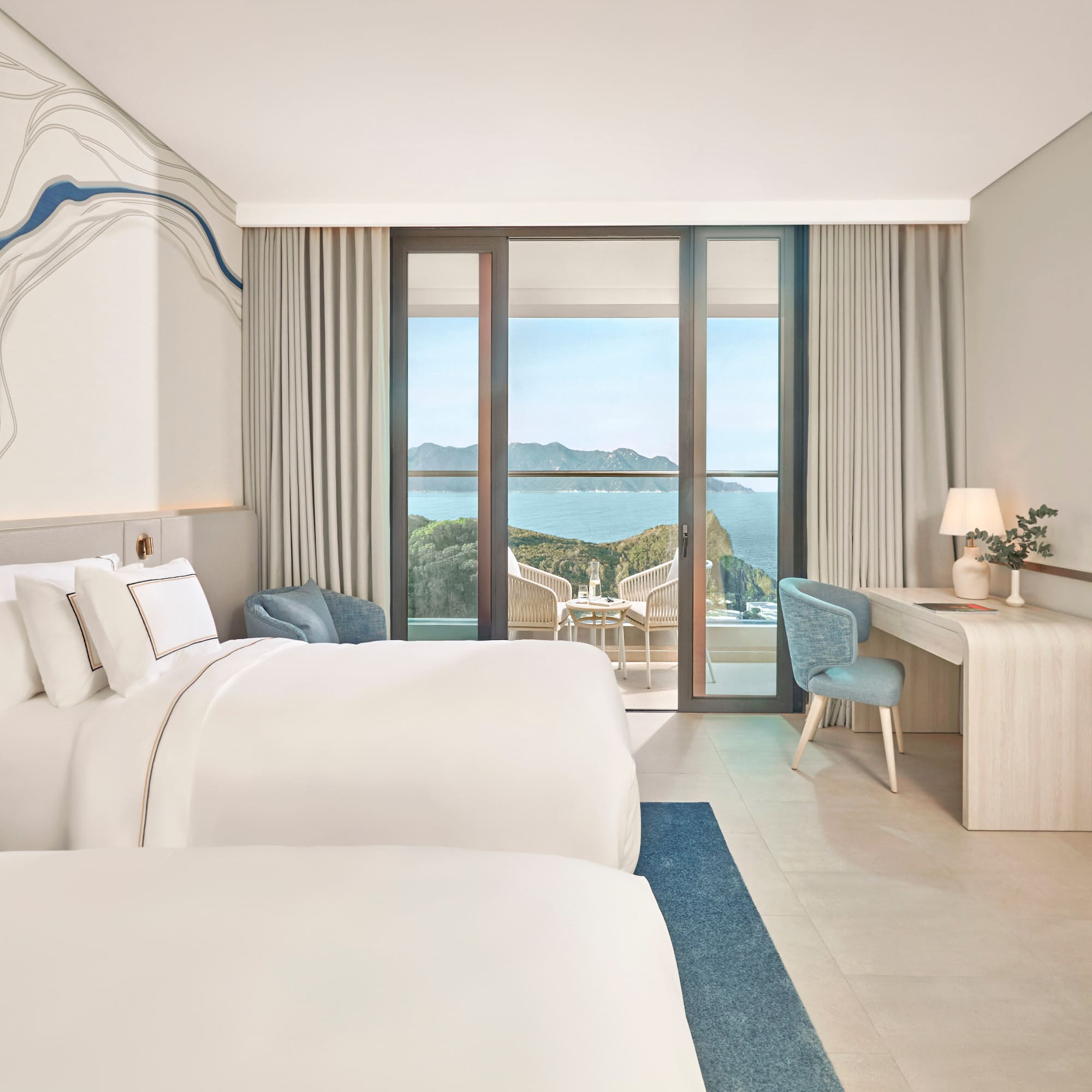 Bright hotel room with two beds, large balcony overlooking ocean and cliffs.