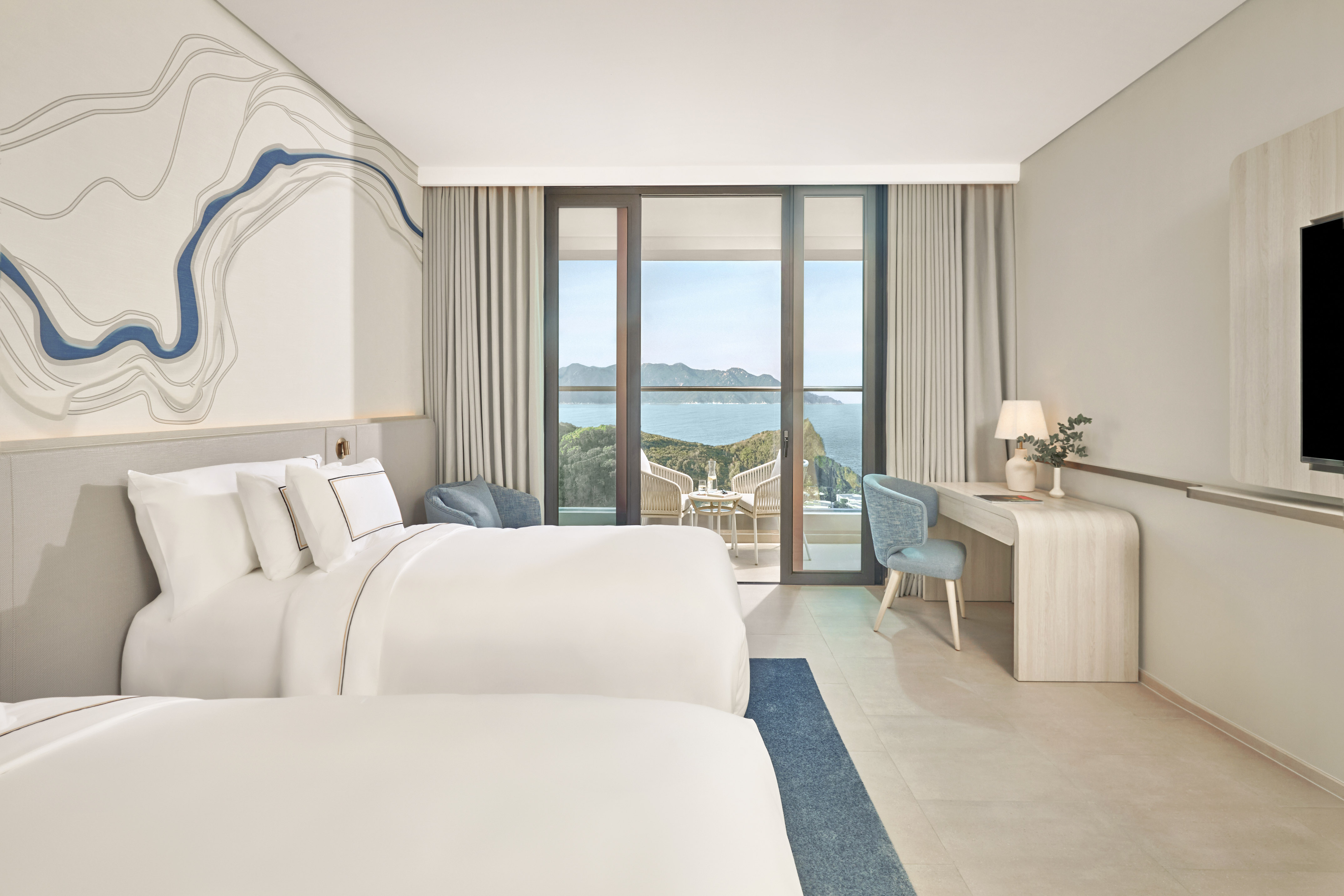 Bright hotel room with two beds, large balcony overlooking ocean and cliffs.
