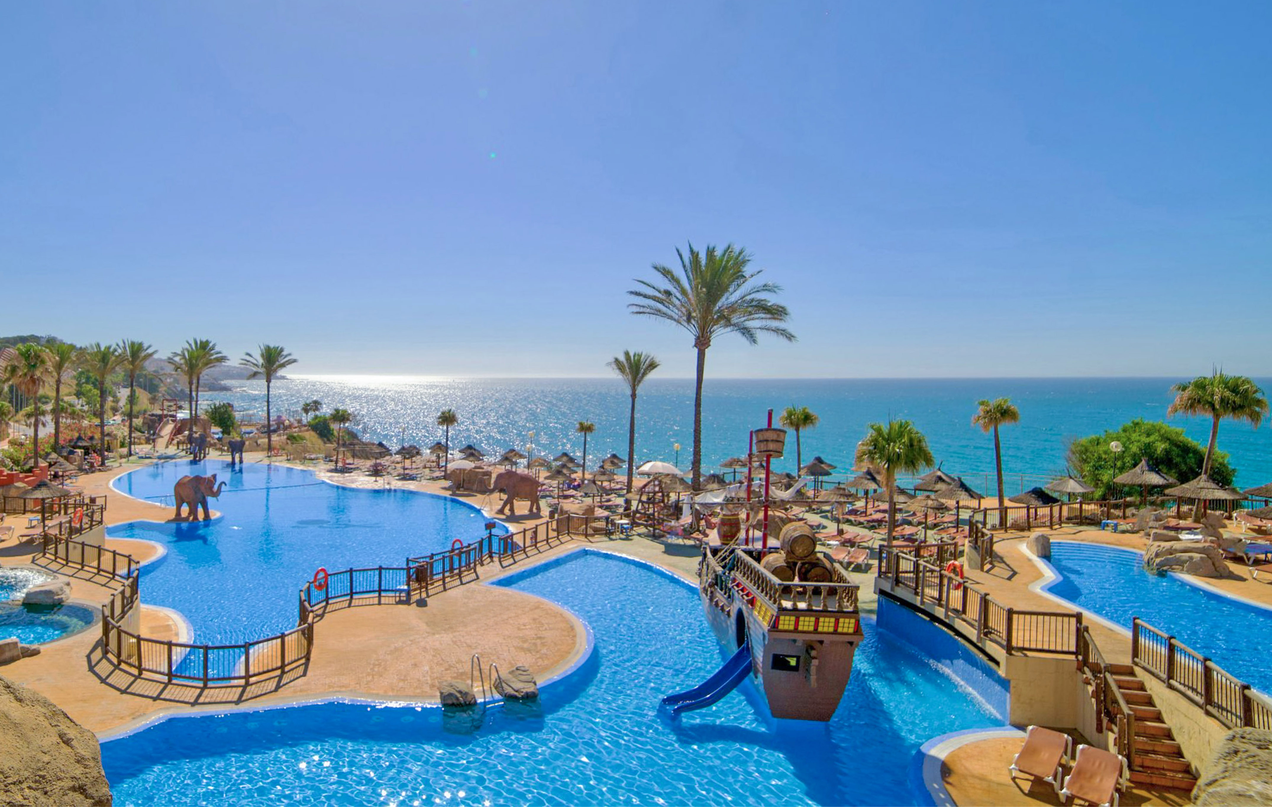 Tropical resort pool with pirate ship, elephant, palm trees, and sea.