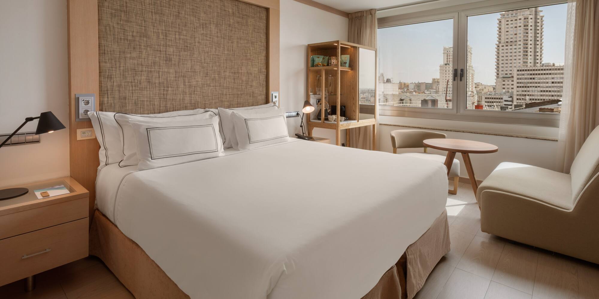 Hotel room with king bed, city view, modern desk, and light wood accents.