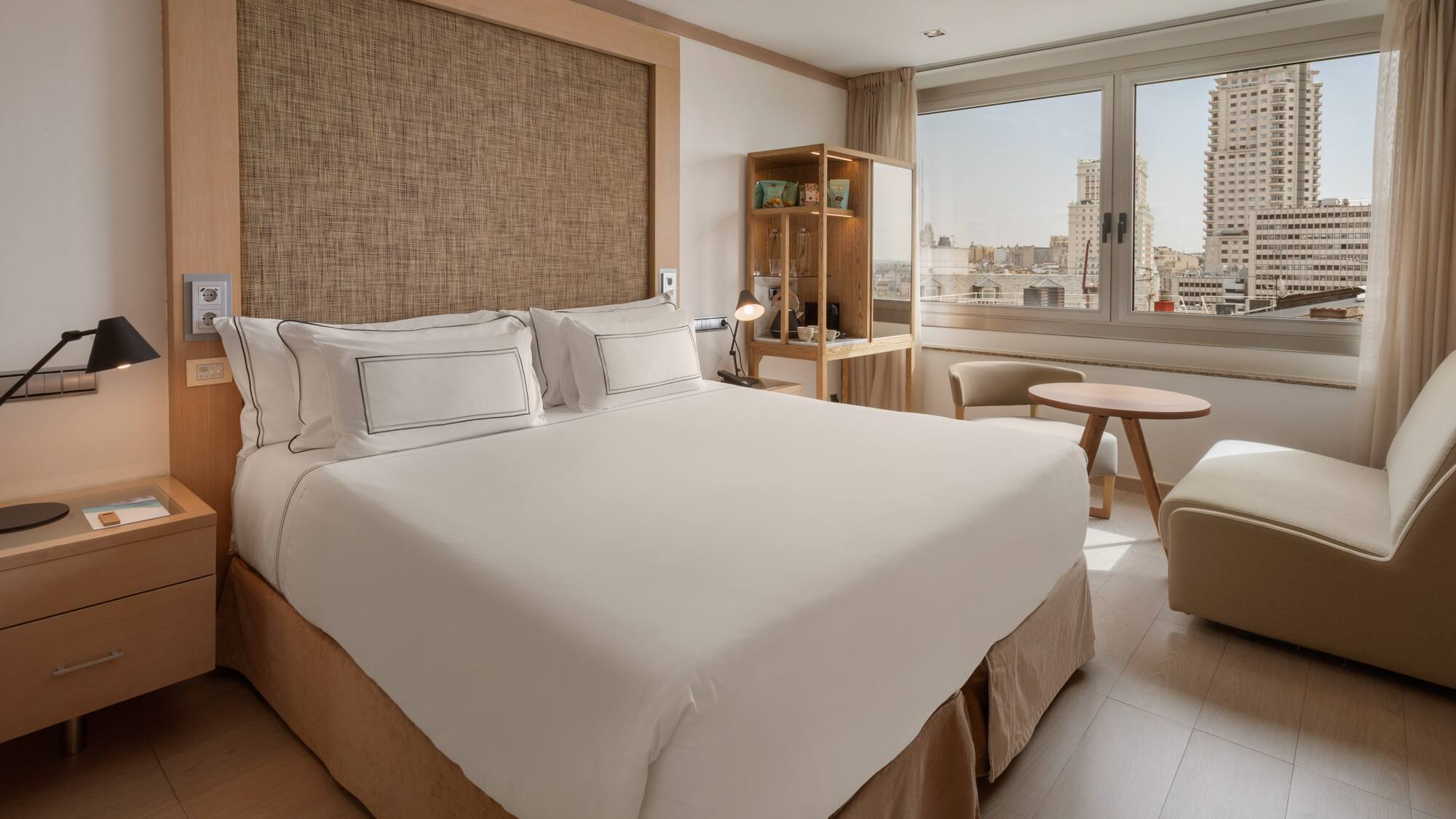 Hotel room with king bed, city view, modern desk, and light wood accents.