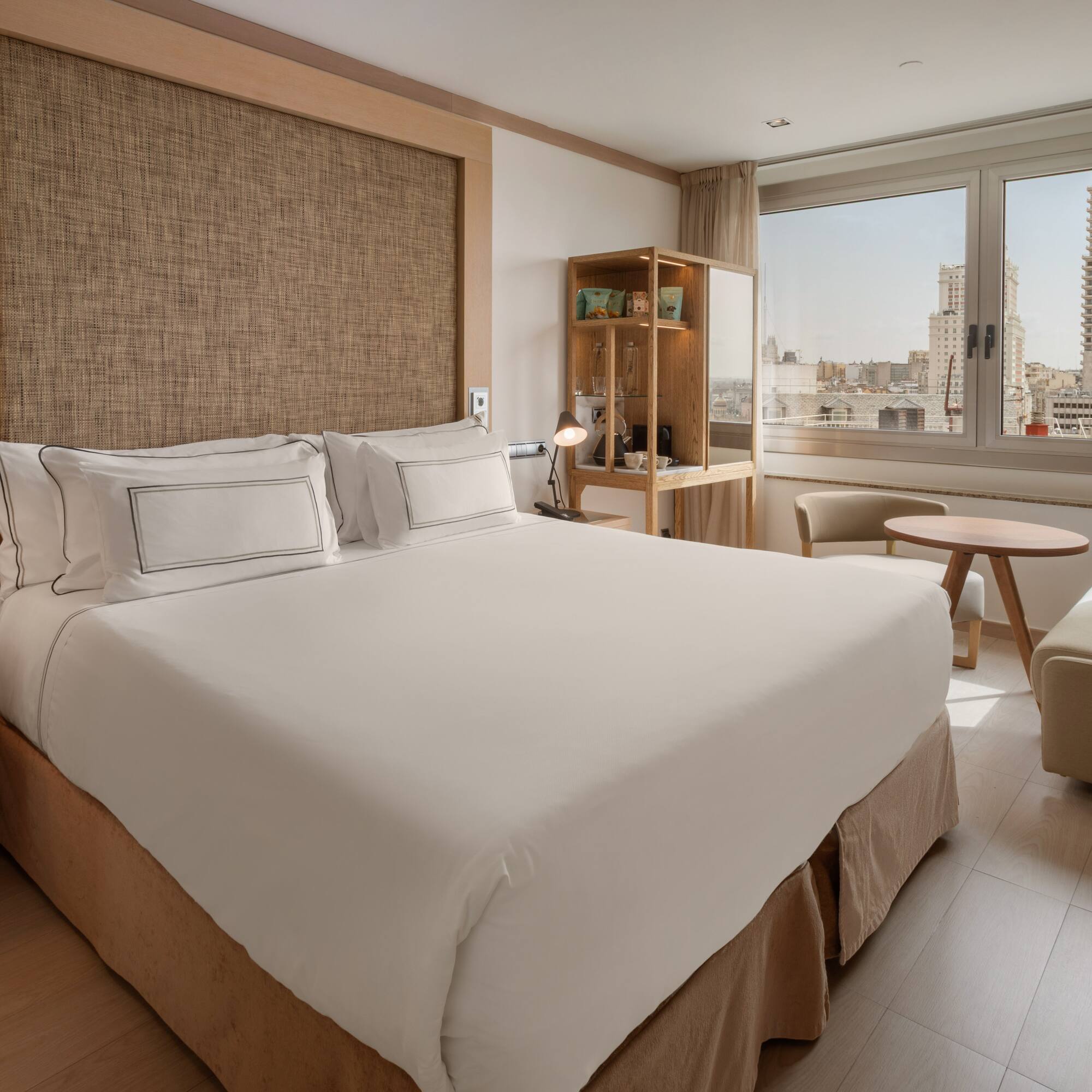 Hotel room with king bed, city view, modern desk, and light wood accents.