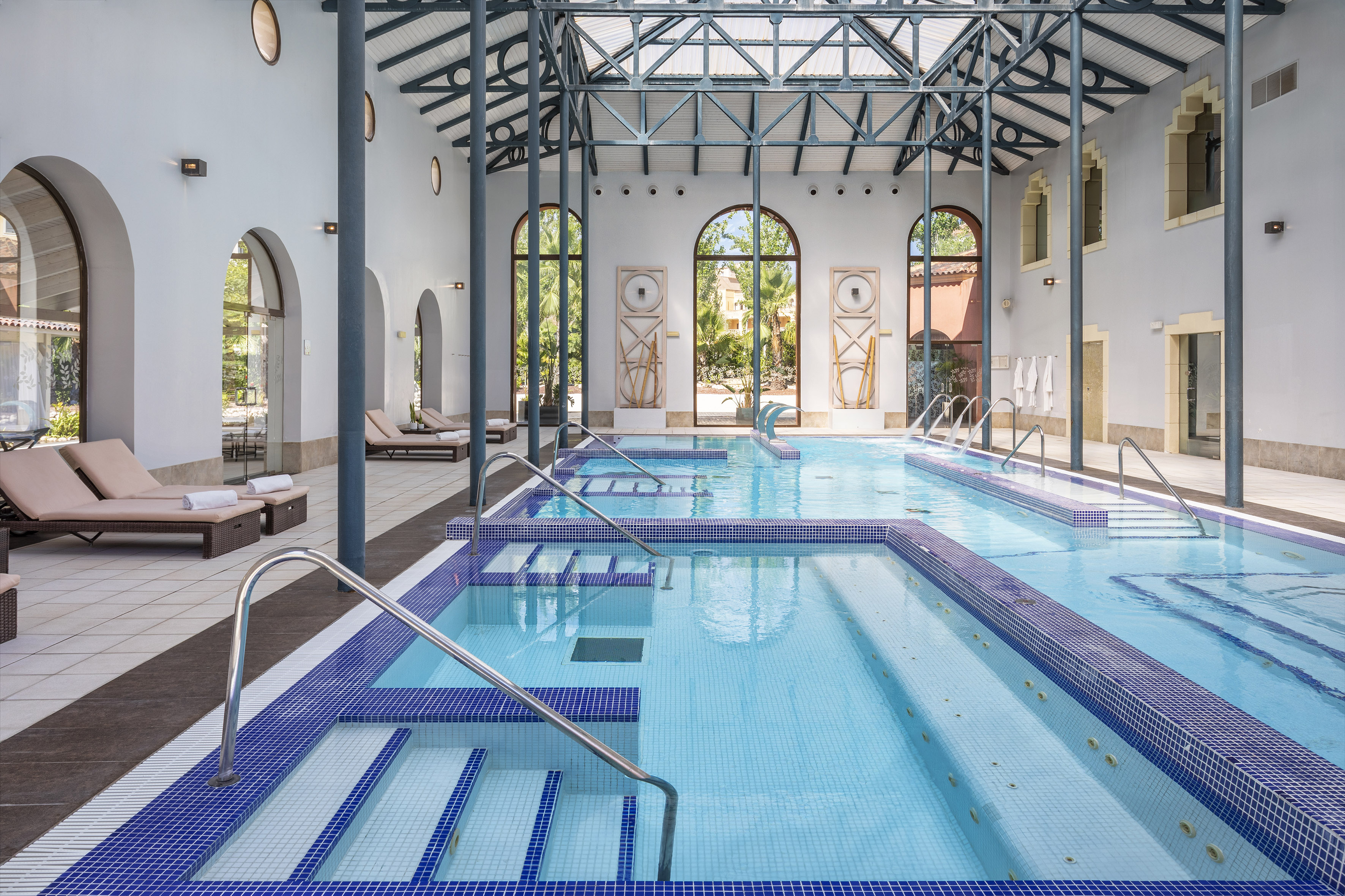 Indoor wellness spa pool with blue mosaic, arched windows, and lounge chairs.