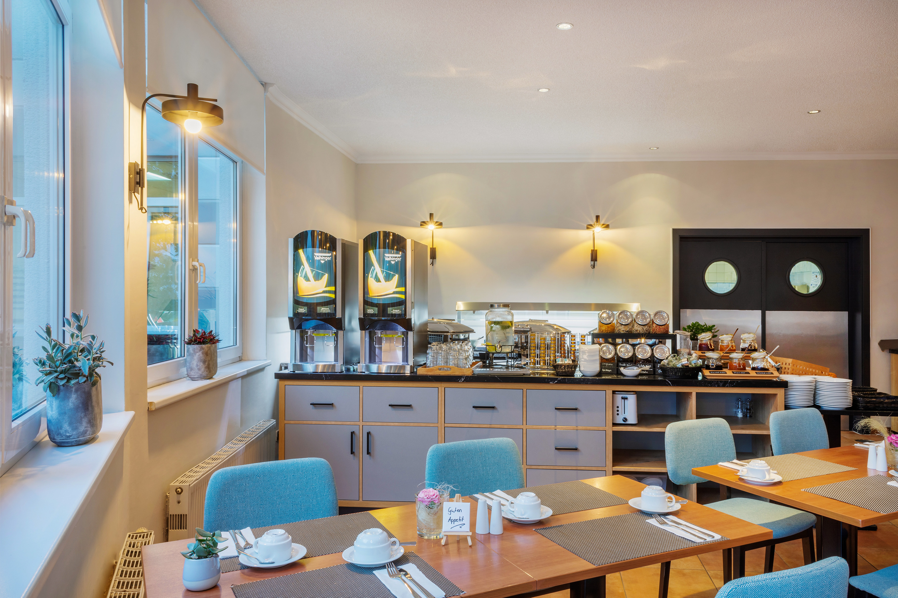 Bright hotel breakfast room with buffet, juice, and set dining tables.