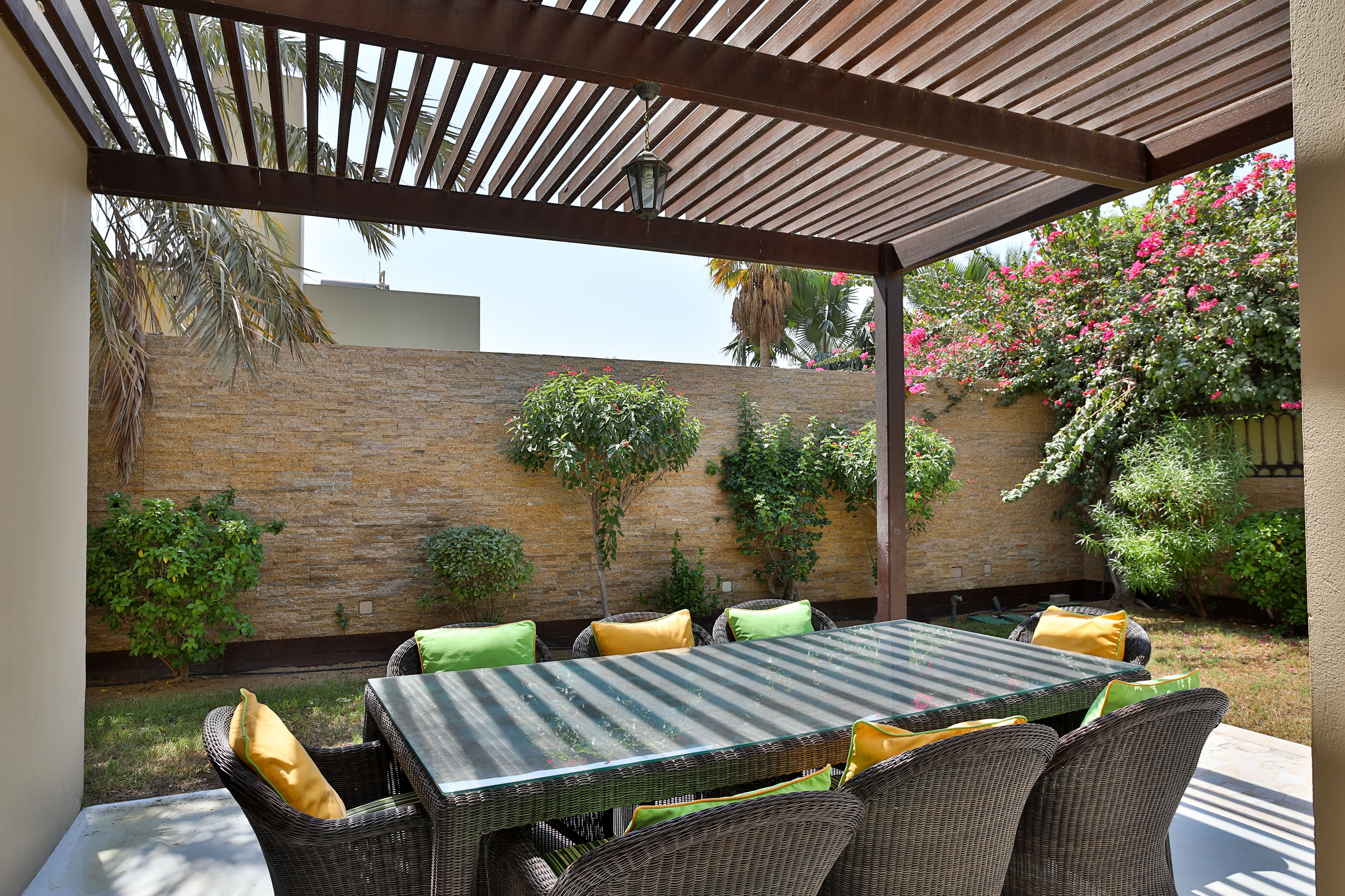 Pergola-covered outdoor dining area with wicker furniture, vibrant cushions, lush garden.