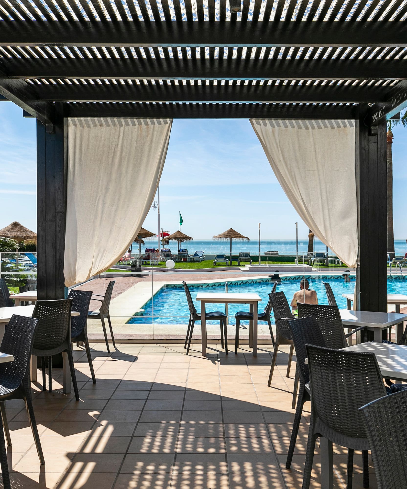 Outdoor restaurant patio with pergola, poolside view of ocean, beach, and palm trees.