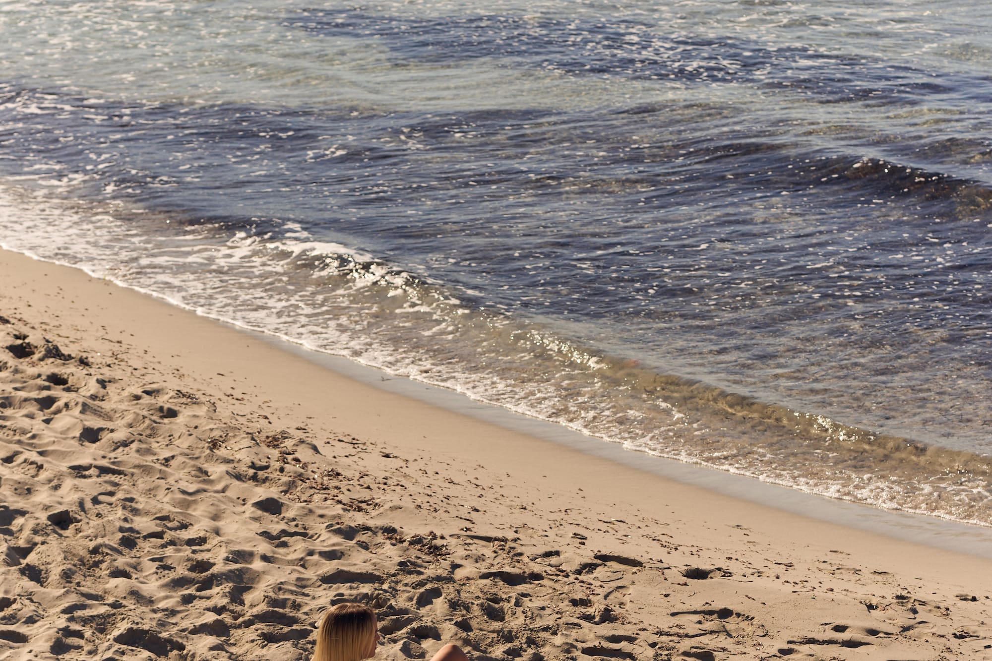 Woman sunbathing on sandy beach by ocean with a straw bag.