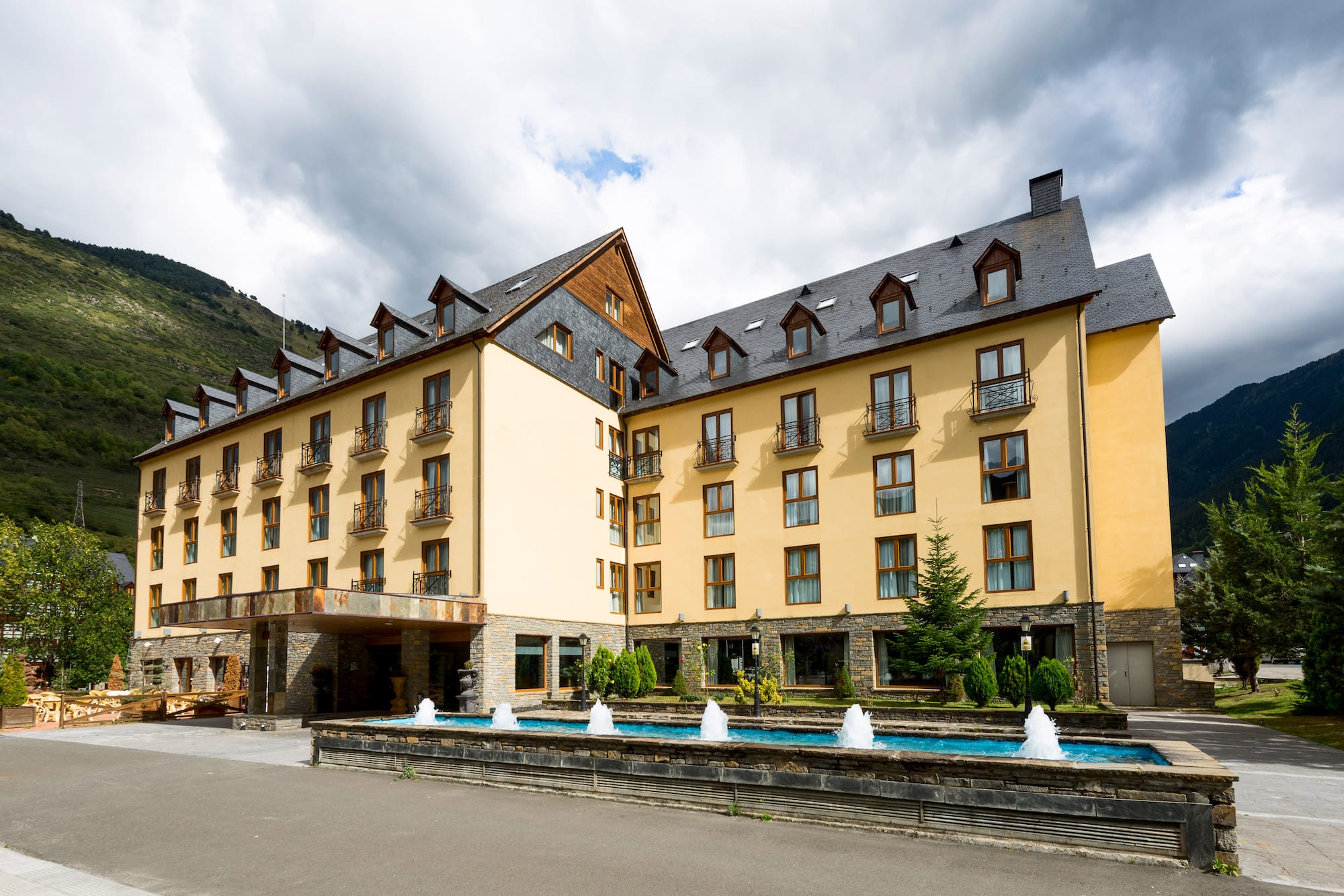 Mountain resort hotel with yellow facade, dark roof, and water fountain.
