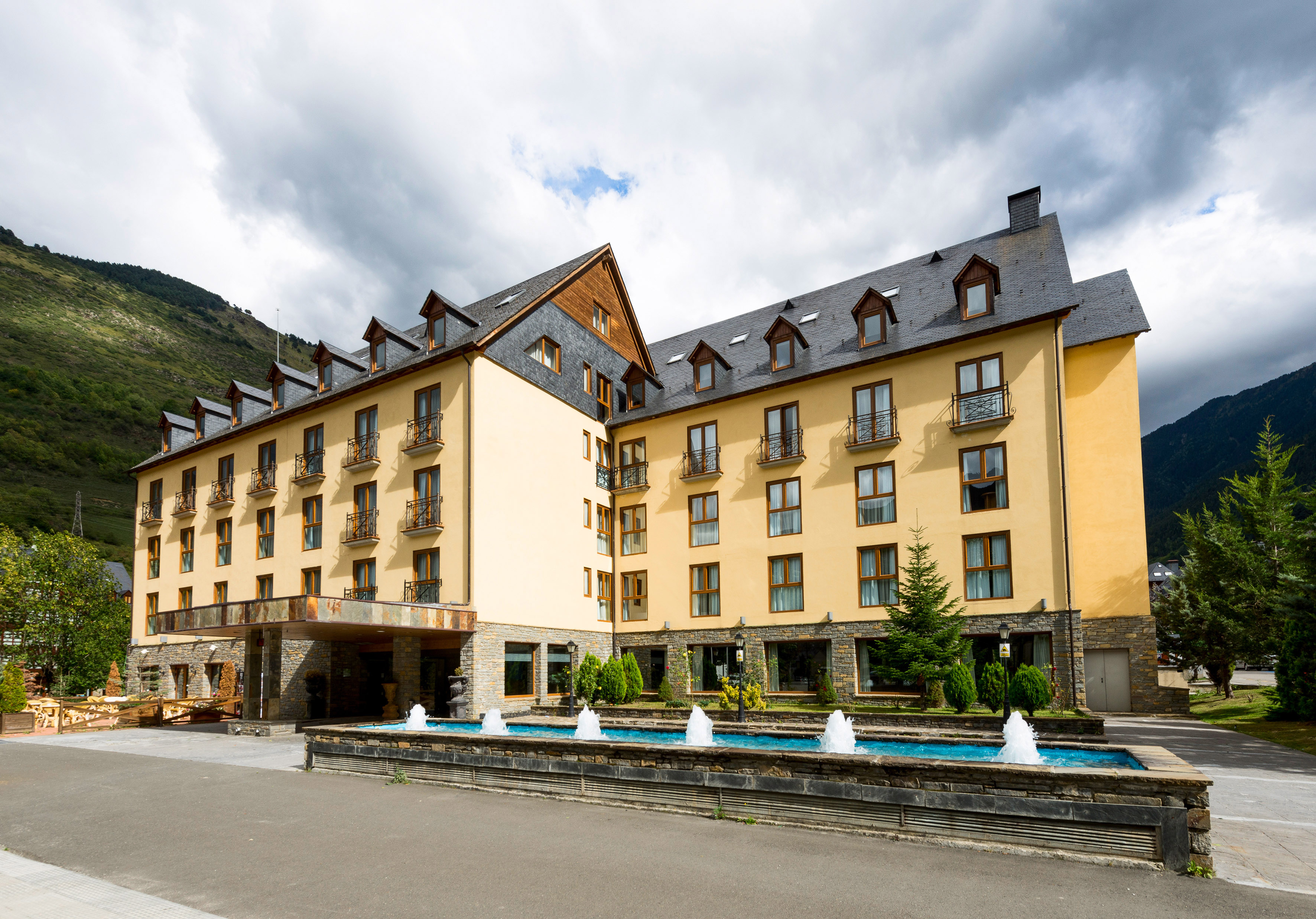 Mountain resort hotel with yellow facade, dark roof, and water fountain.