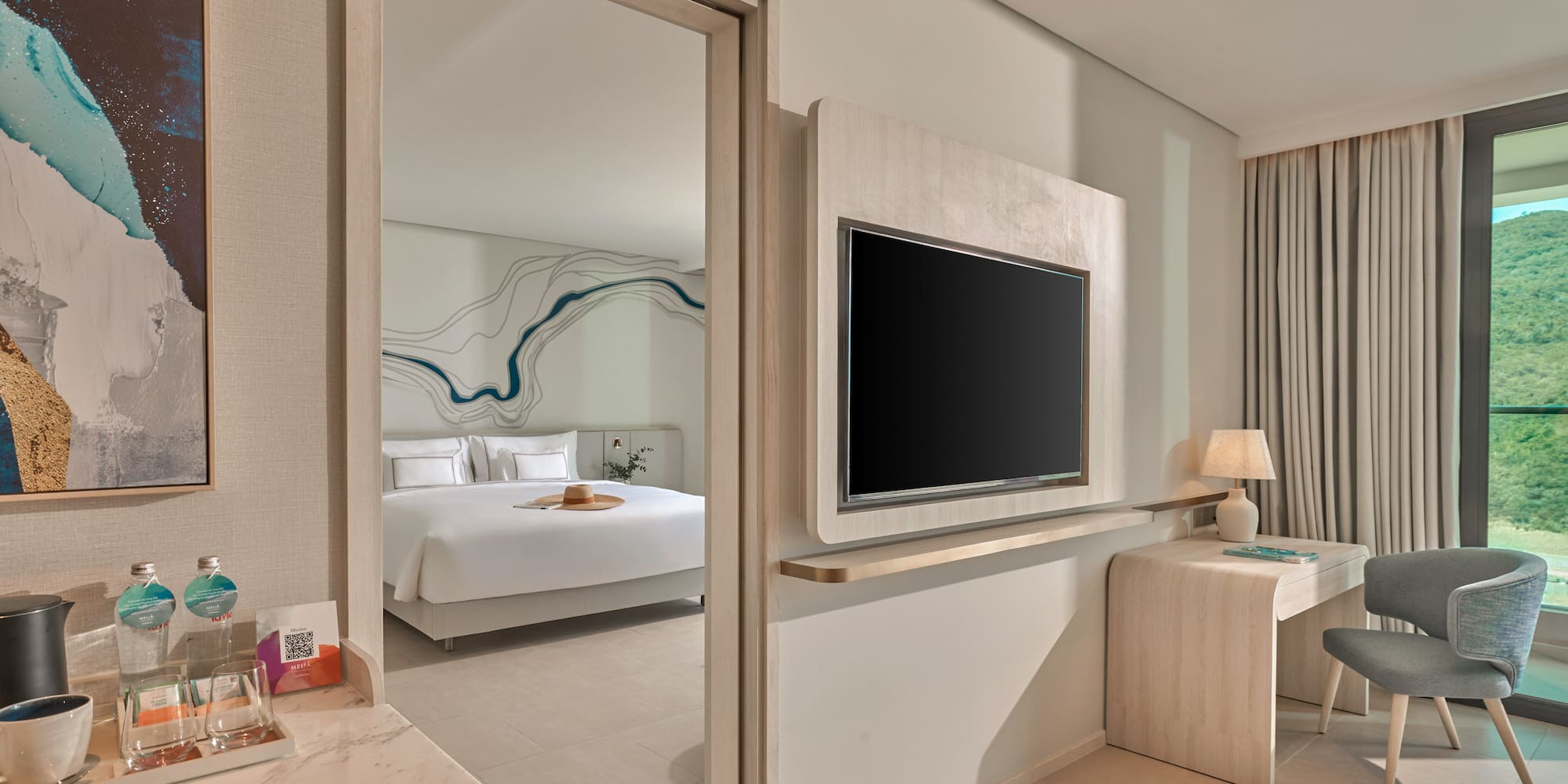 Modern hotel suite with open bedroom, TV, desk, and scenic mountain view.