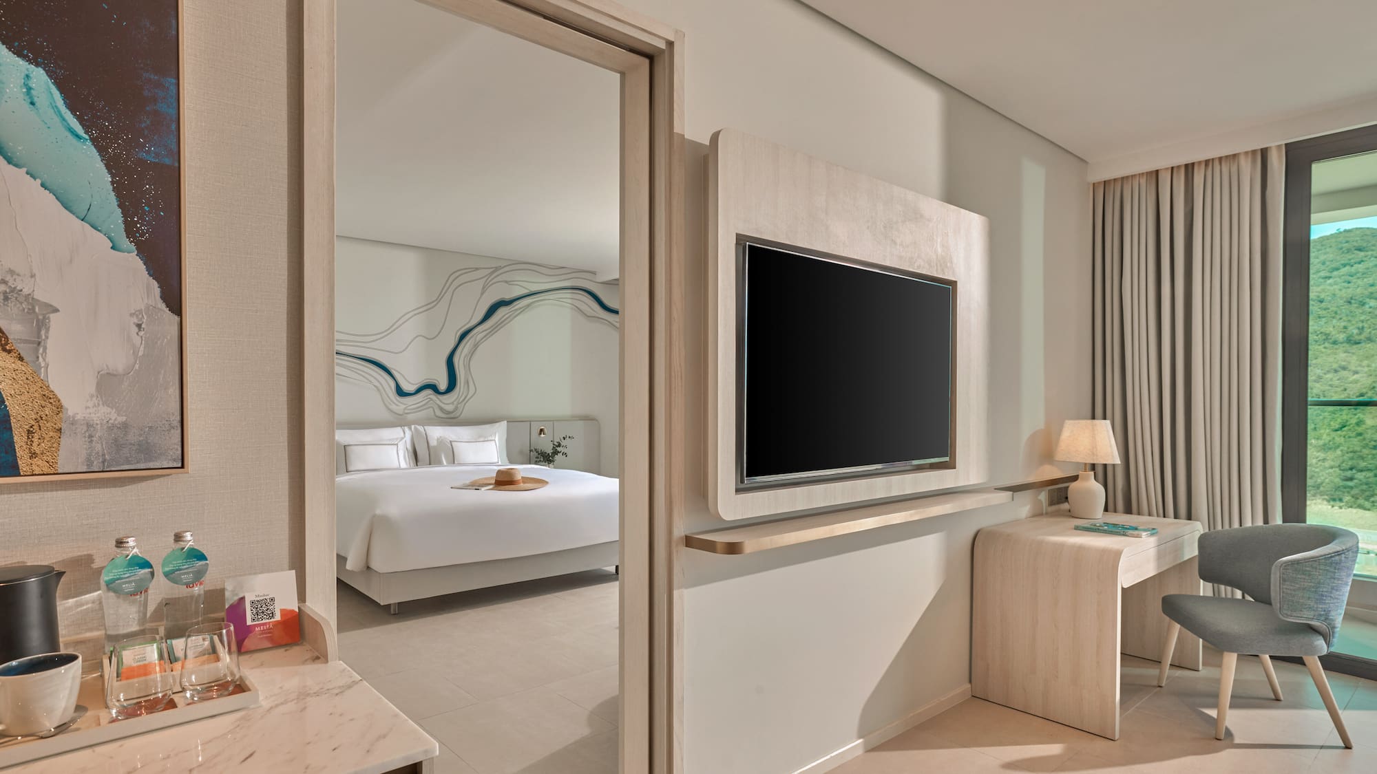 Modern hotel suite with open bedroom, TV, desk, and scenic mountain view.