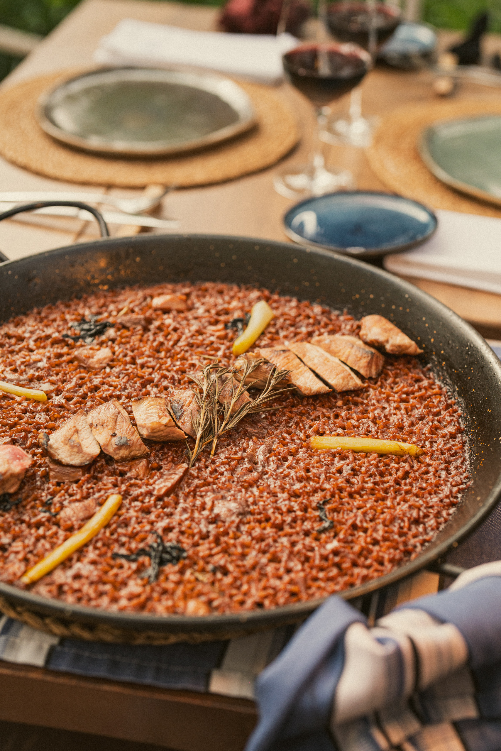 Paella with meat, rosemary, and lemon at an outdoor dining setting.