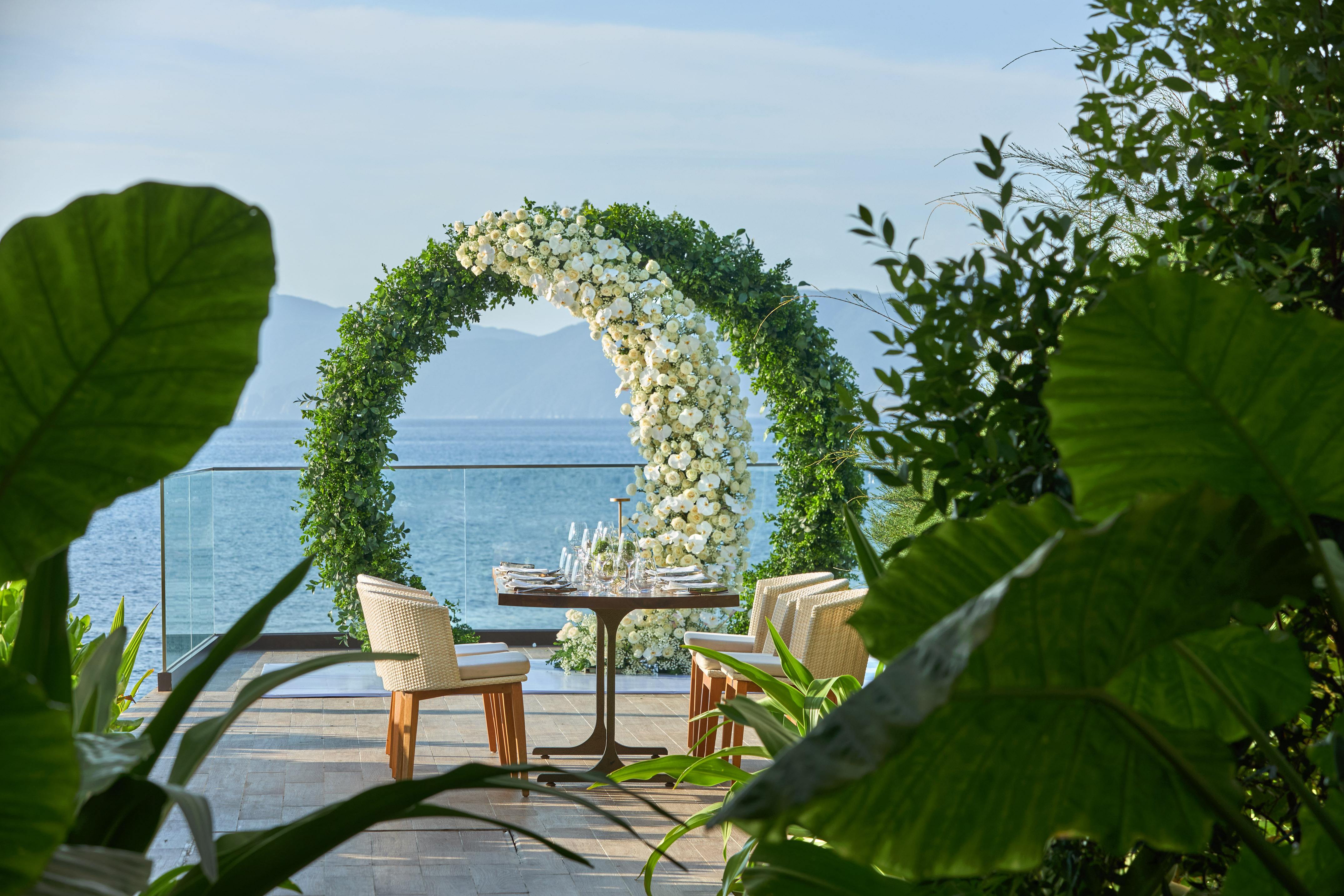 Waterfront dining with white rose arch, set table for two, ocean backdrop.