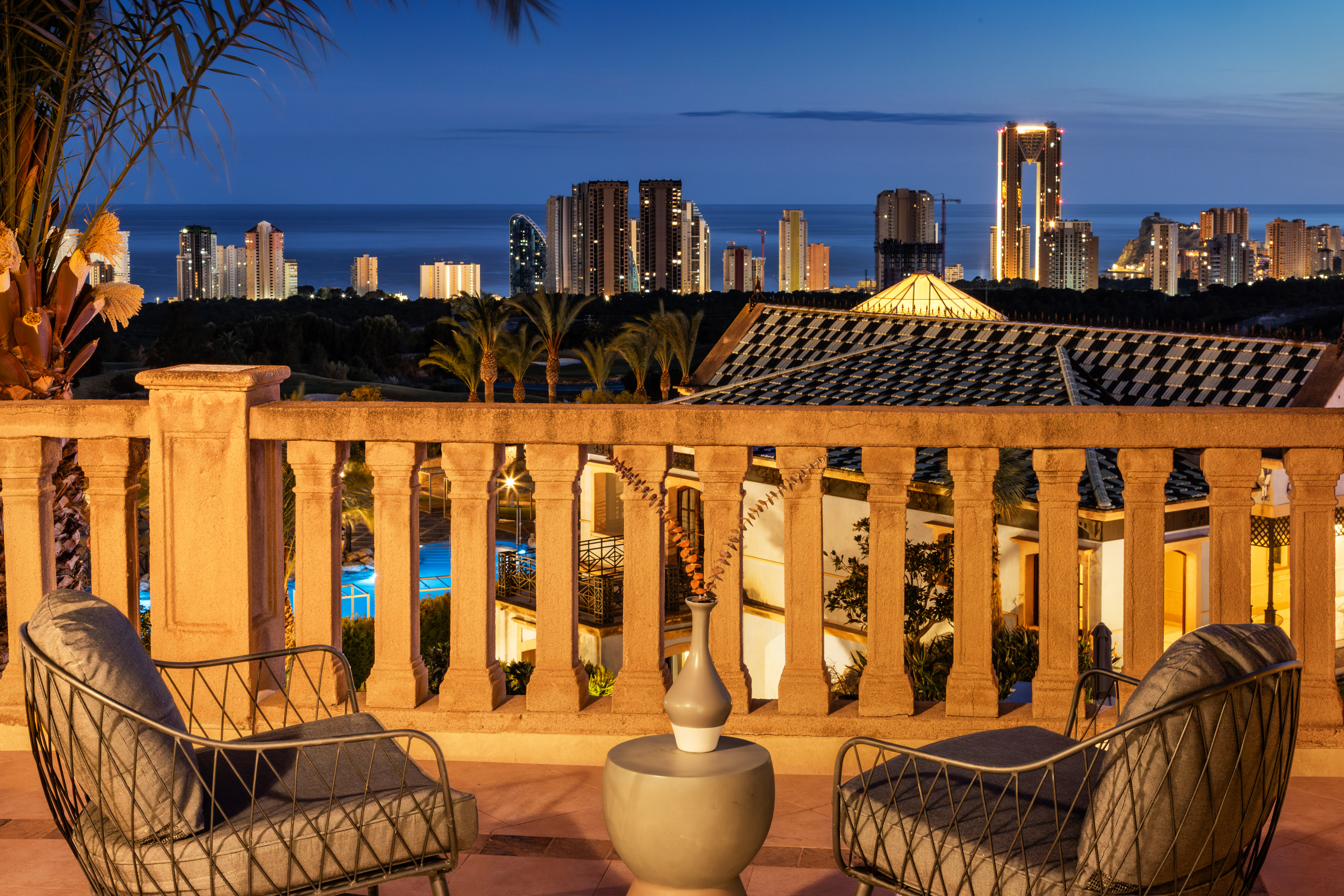 Terrace view of Benidorm city skyline, ocean, and palm trees at dusk.
