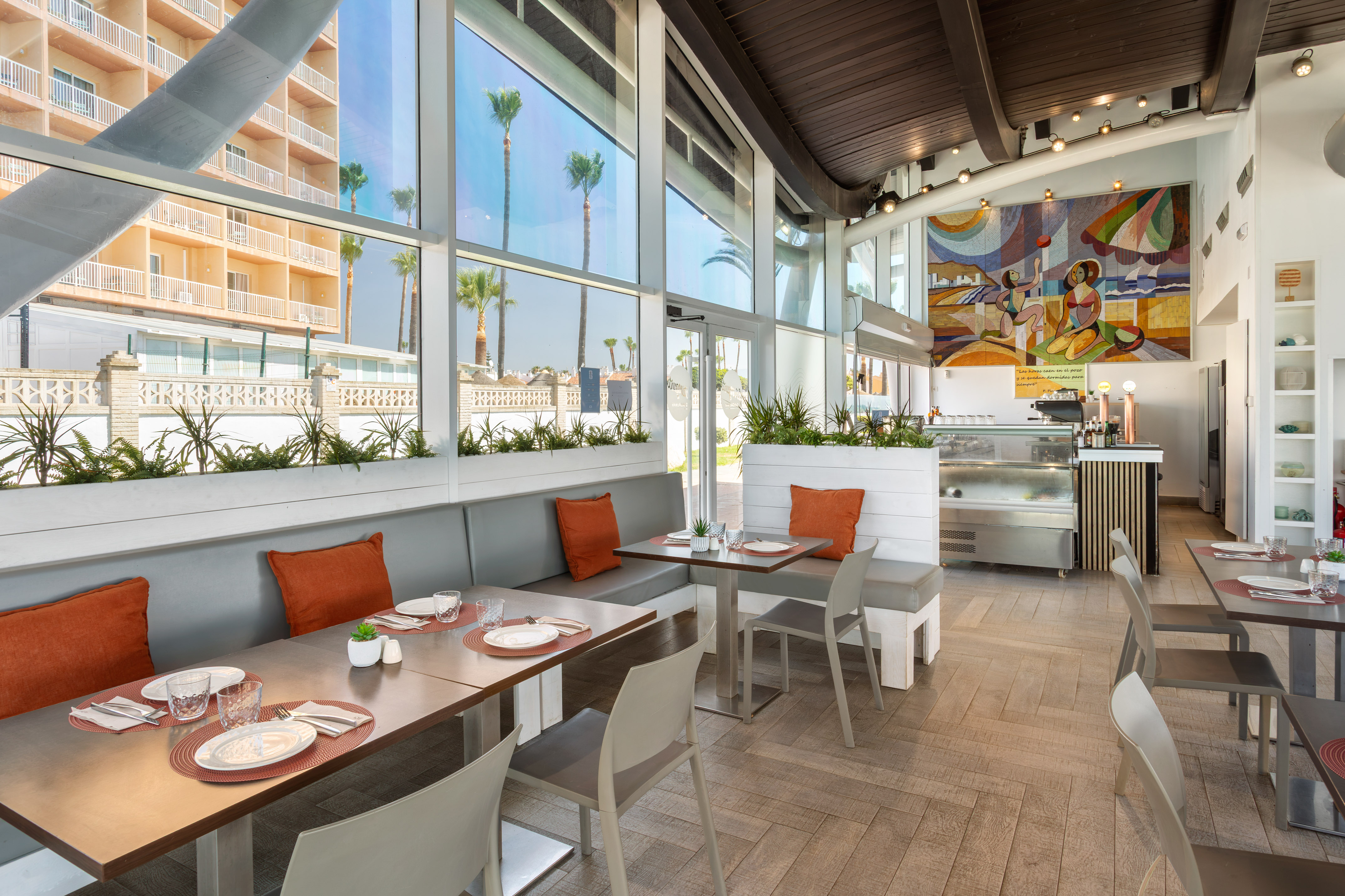 Interior de restaurante moderno con grandes ventanales, palmeras y mural vibrante.
