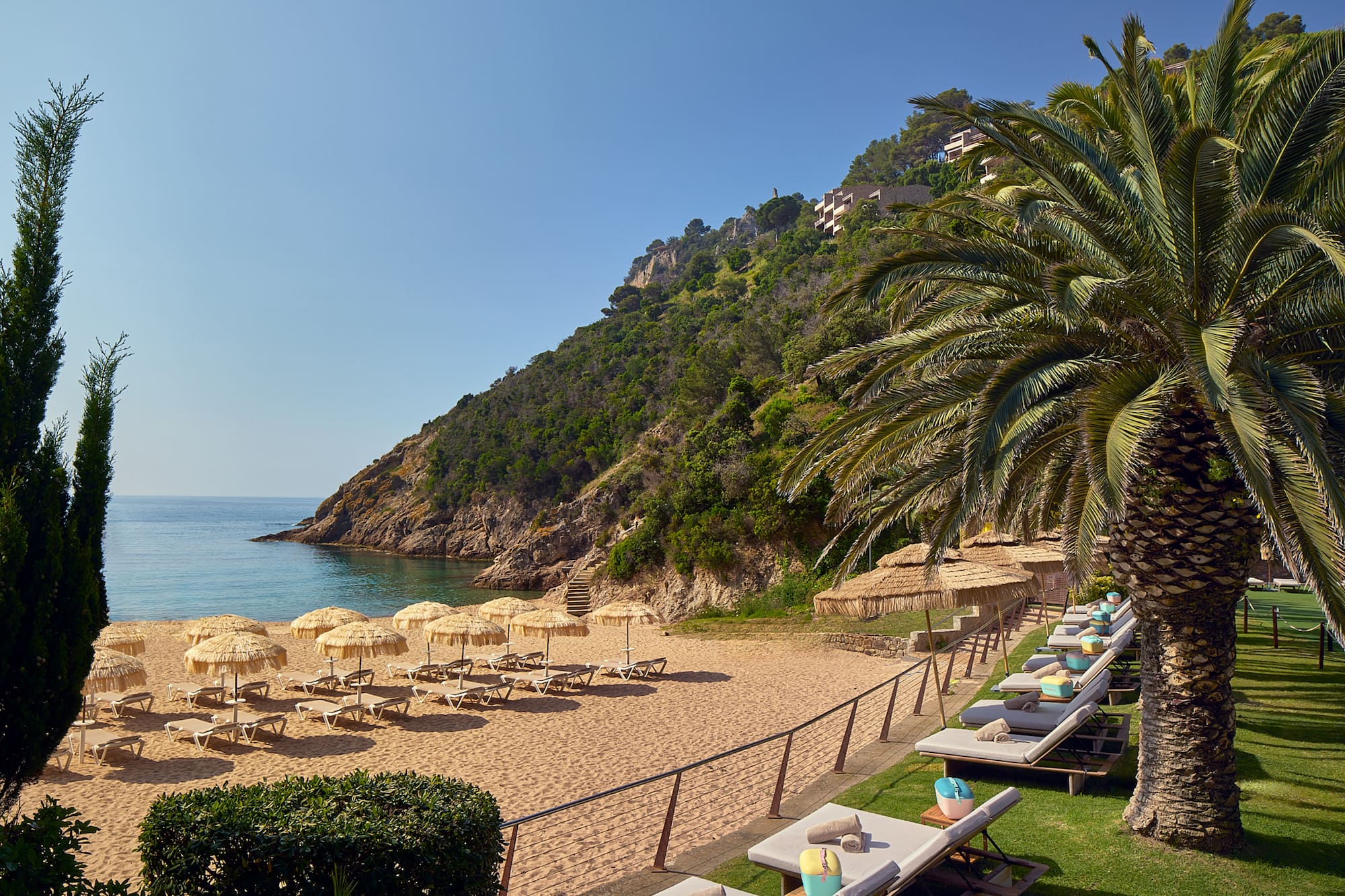 Resort beach with straw umbrellas, loungers, calm sea, and green hills.