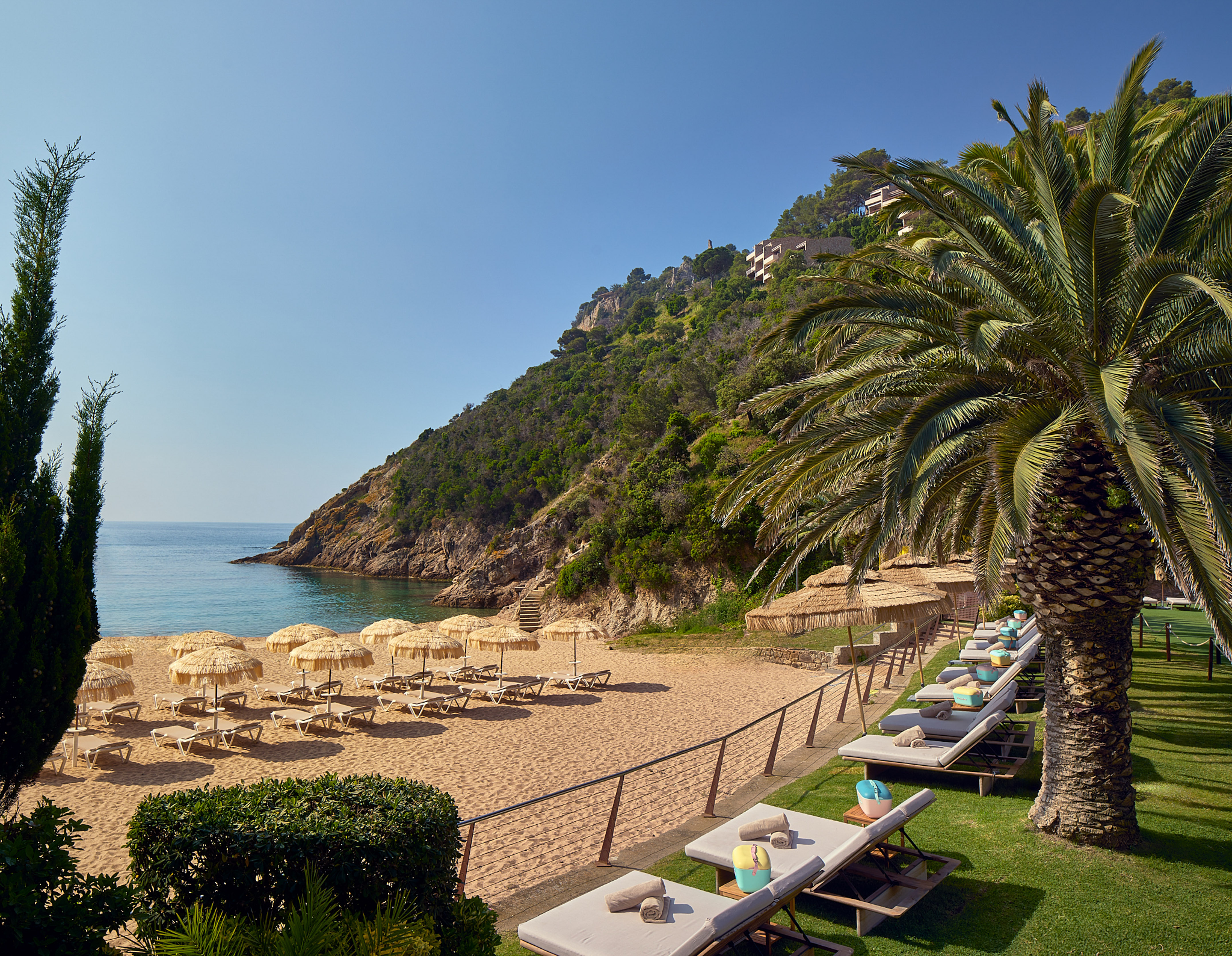 Resort beach with straw umbrellas, loungers, calm sea, and green hills.