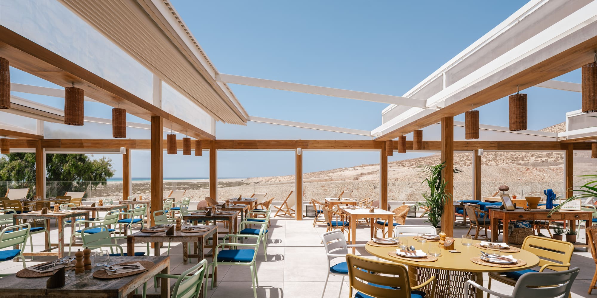 Coastal outdoor restaurant with modern tables, colorful chairs, and sandy beach views.