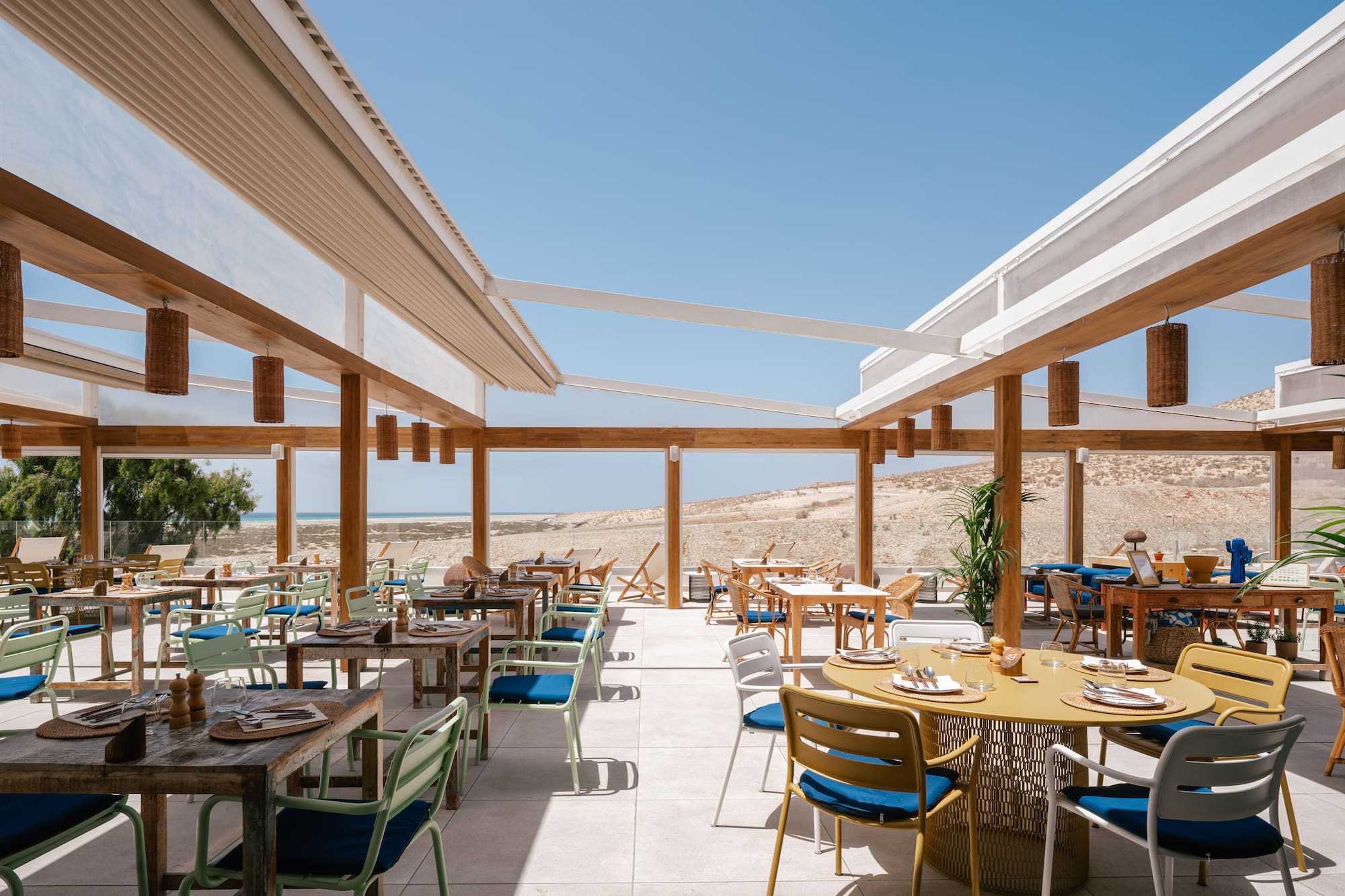 Coastal outdoor restaurant with modern tables, colorful chairs, and sandy beach views.