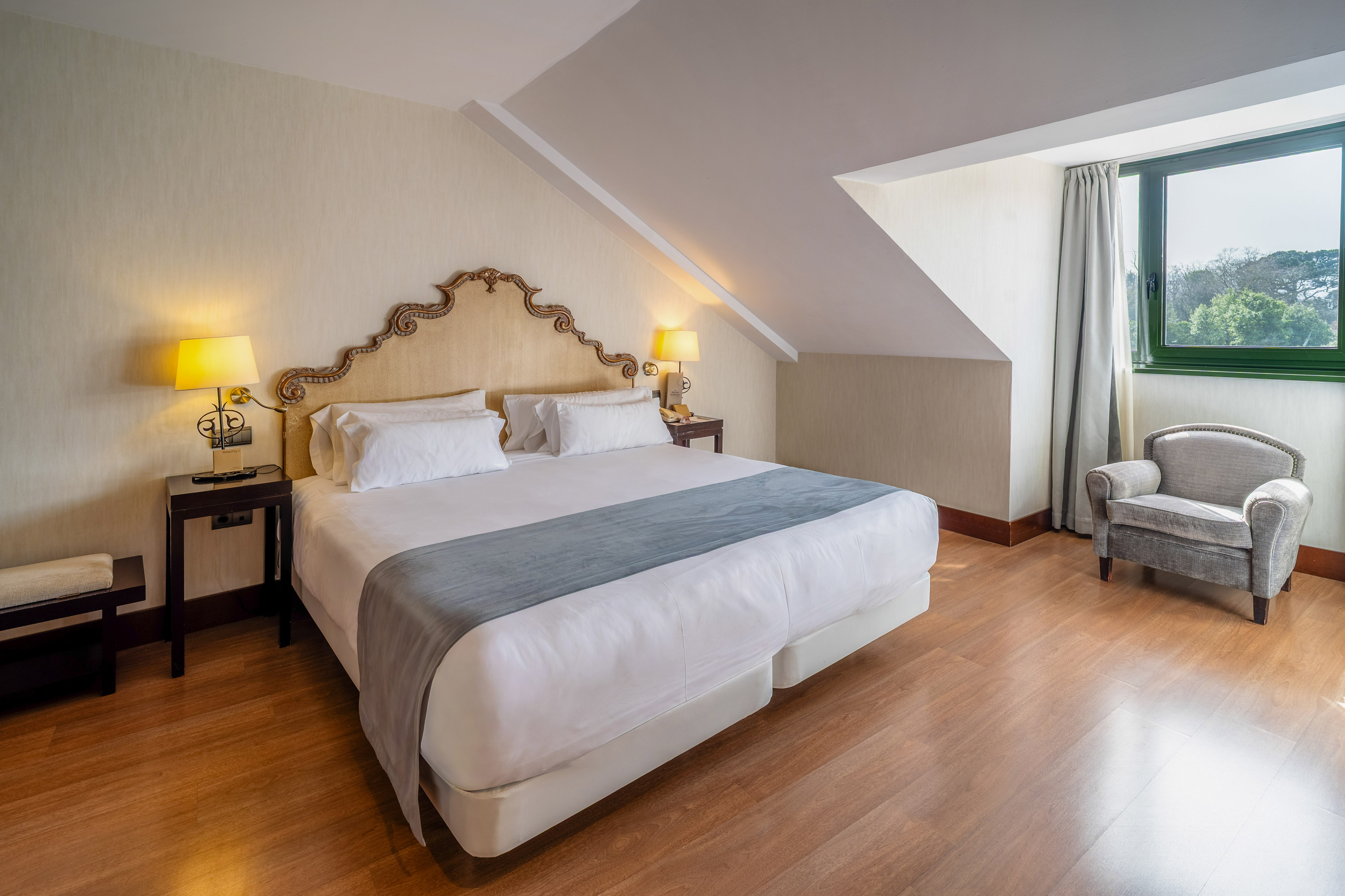 Hotel bedroom with king bed, sloped ceiling, ornate headboard, armchair, and window view.