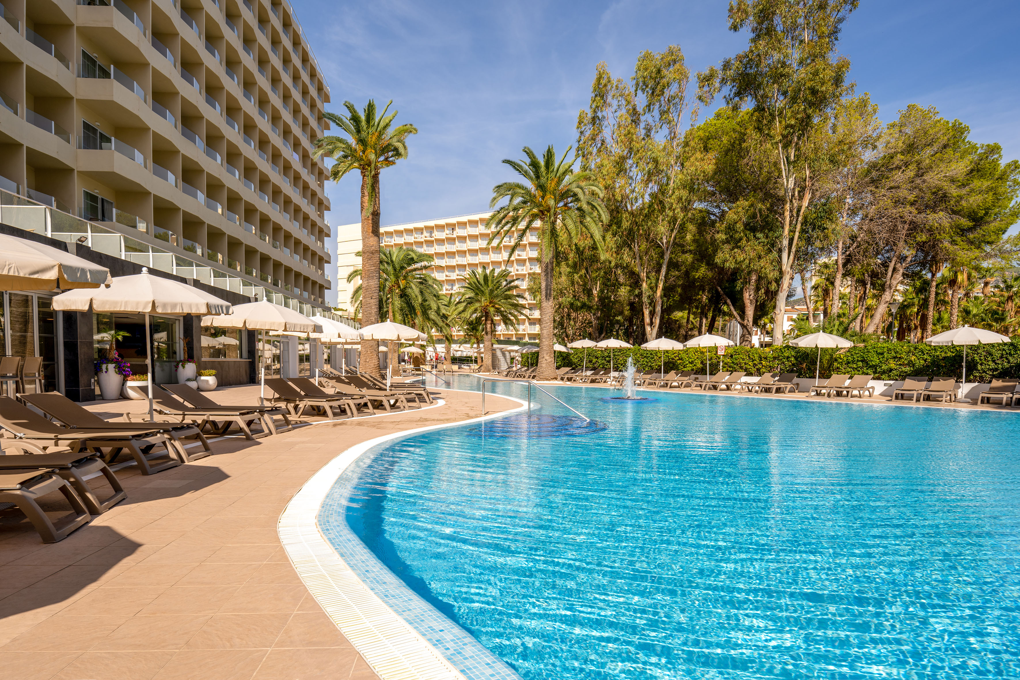 Curved resort swimming pool, palm trees, sun loungers, and hotel buildings.