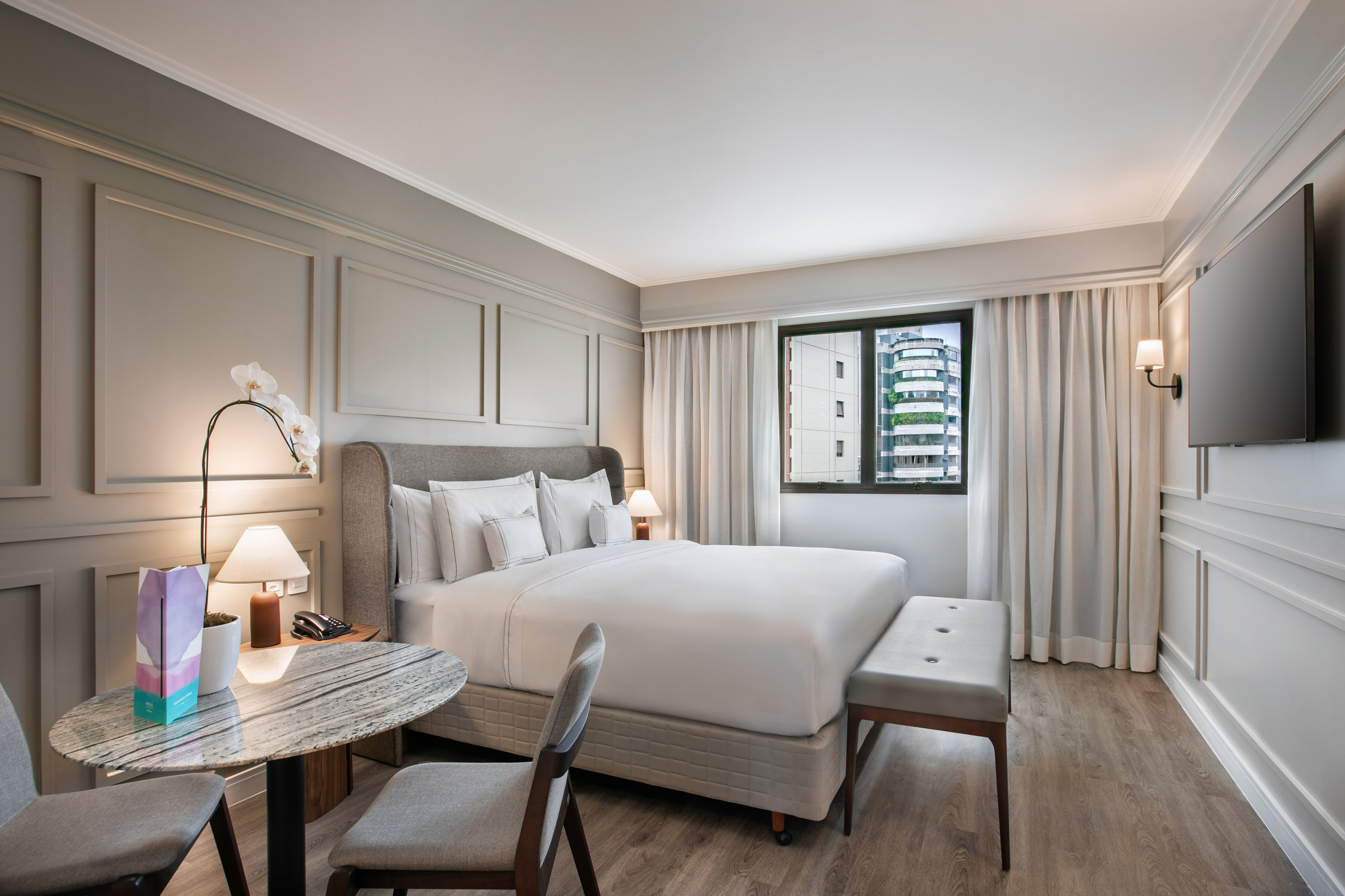 Modern hotel room with king bed, city view, wood floor, and marble table.