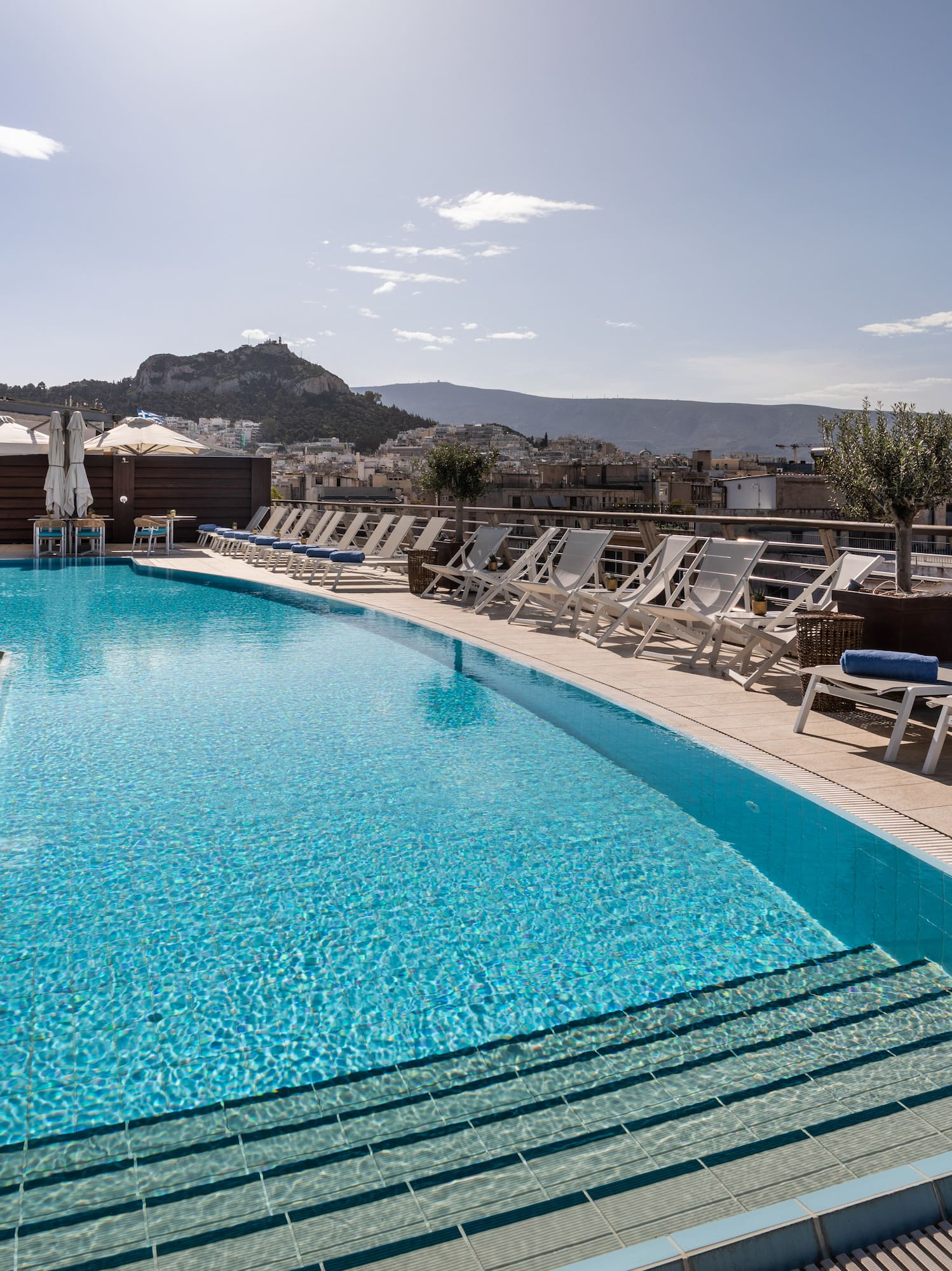 Rooftop pool in Athens with cityscape, sun loungers, and Acropolis view.