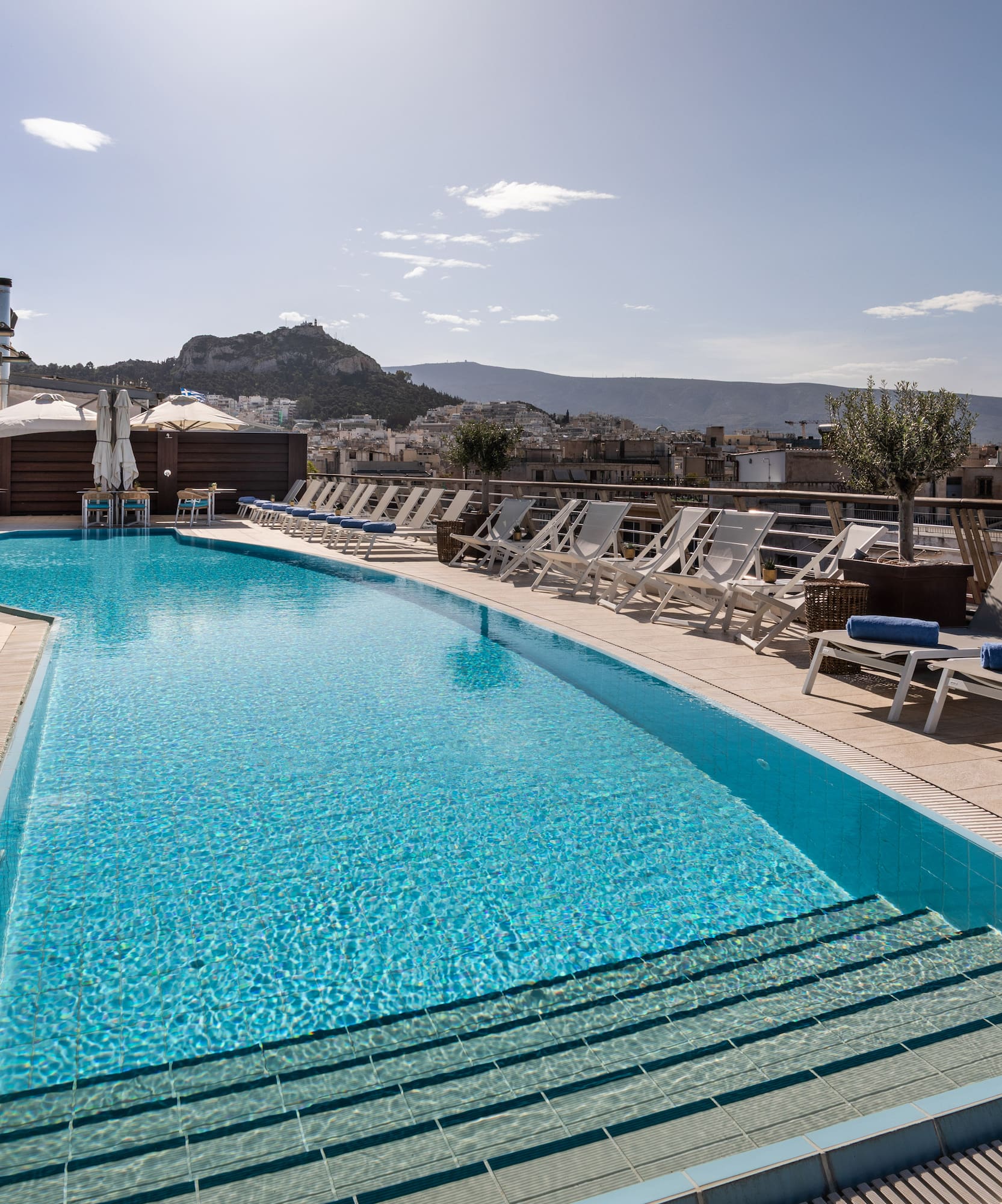 Rooftop pool in Athens with cityscape, sun loungers, and Acropolis view.