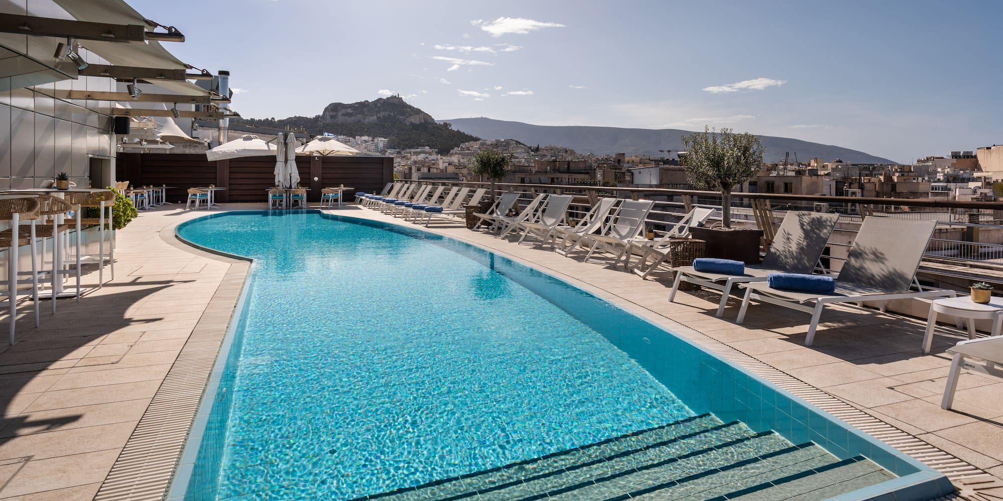 Rooftop pool with sun loungers, bar, and panoramic Athens Acropolis view.