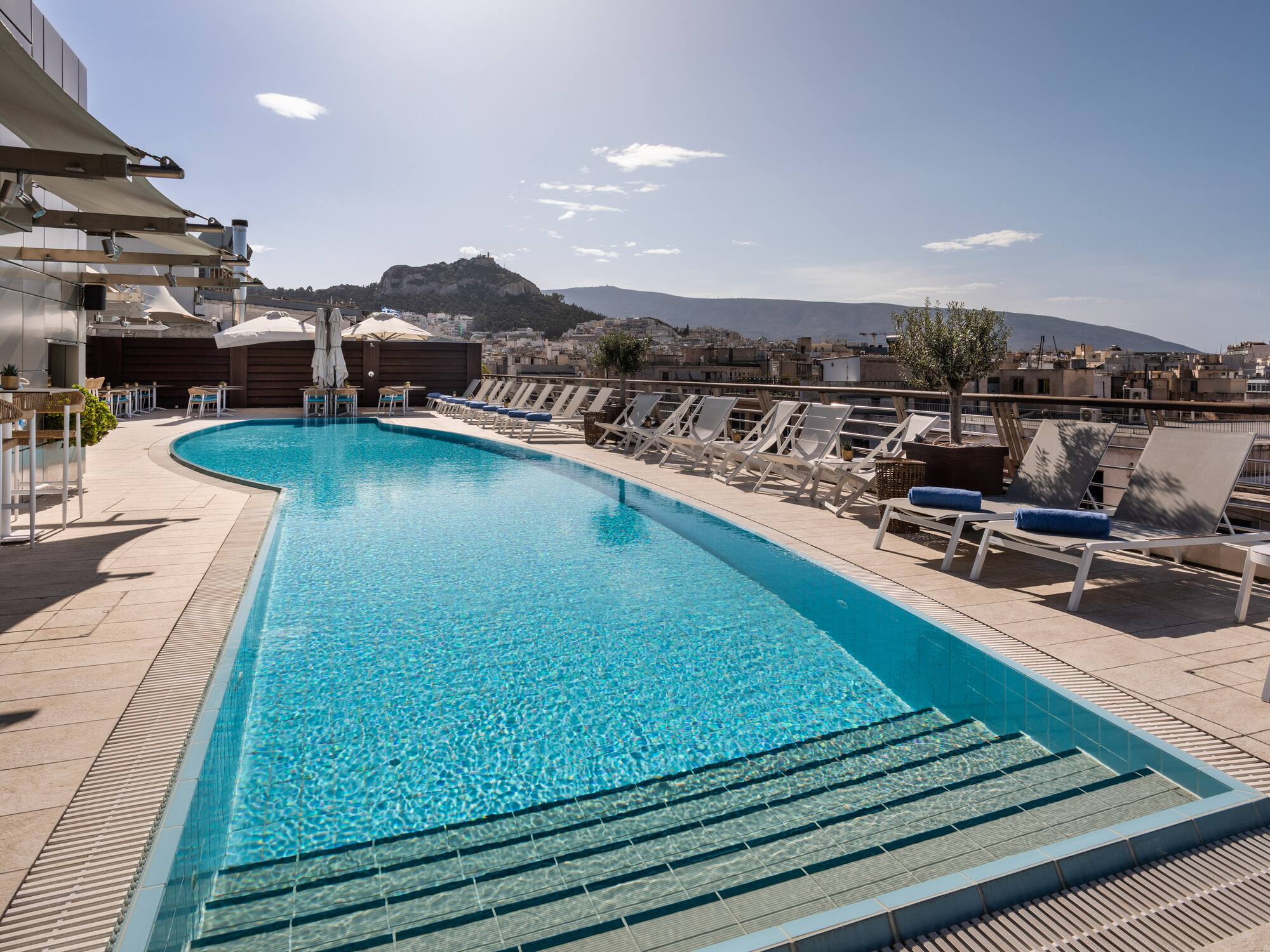 Sunny rooftop pool in Athens with city views, sun loungers, and Acropolis.